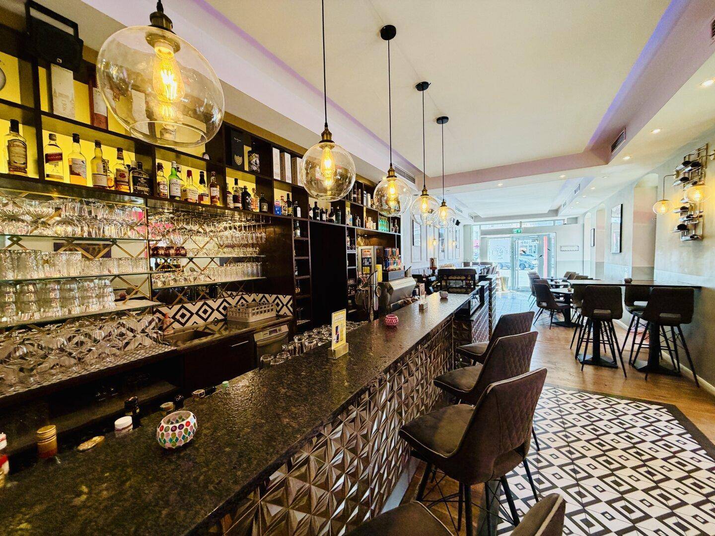 Eine moderne Bar mit dunkler Theke, vielen Gläsern und Flaschen sowie einem stilvollen Bodenmuster.