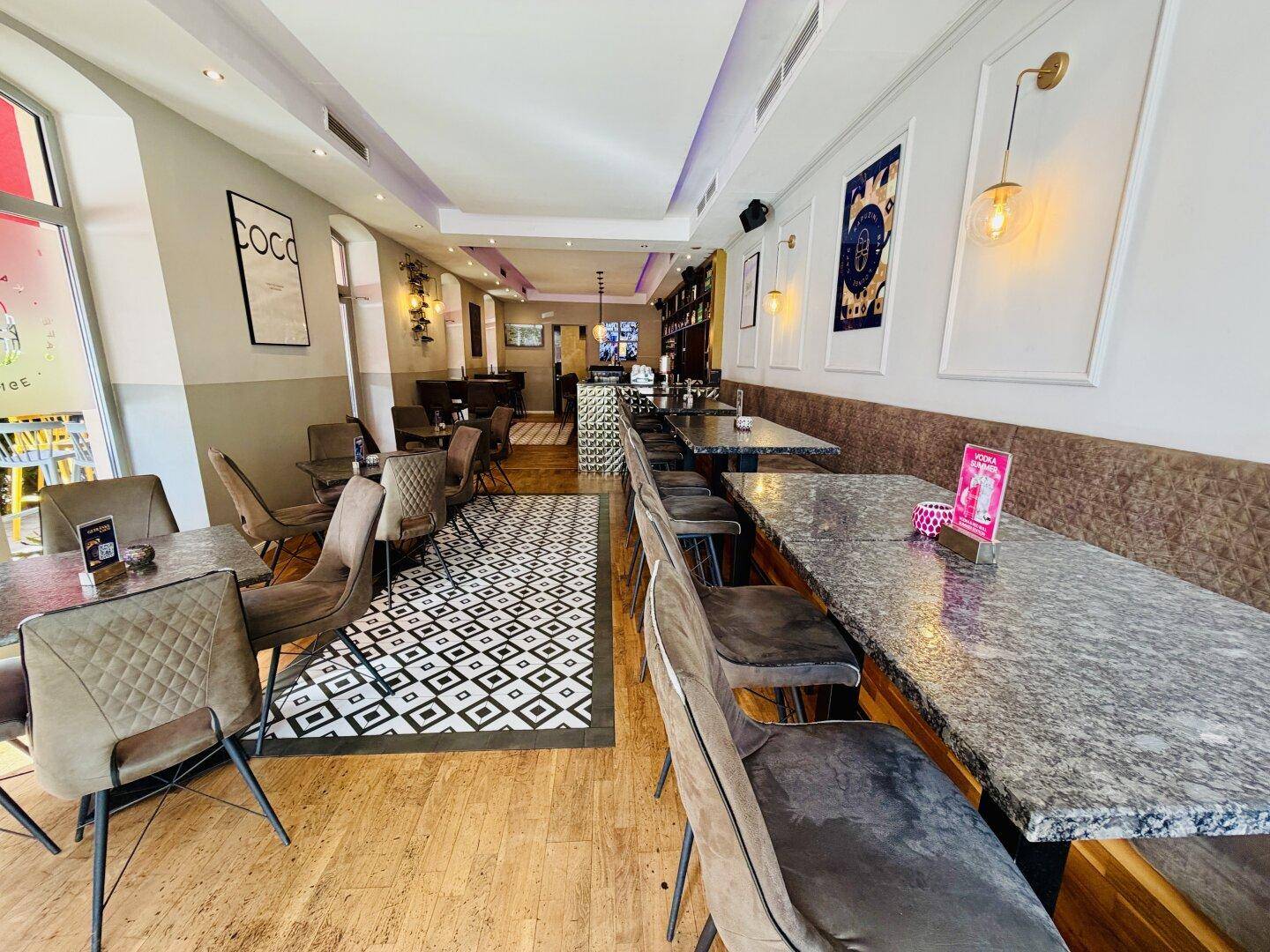 Heller Café-Innenraum mit Holzboden, modernen Tischen und Stühlen sowie großen Fenstern.