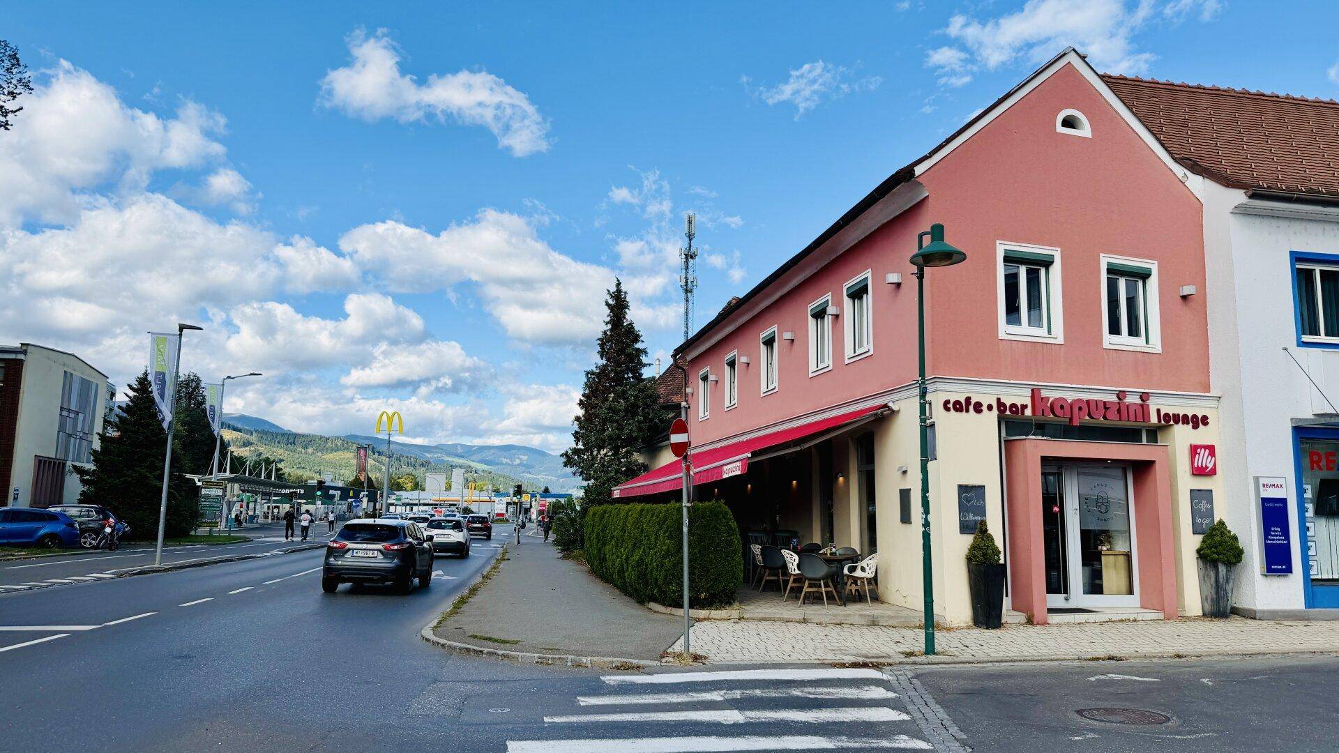Gebäude mit Café im Erdgeschoss an einer belebten Straße mit Bergen im Hintergrund.