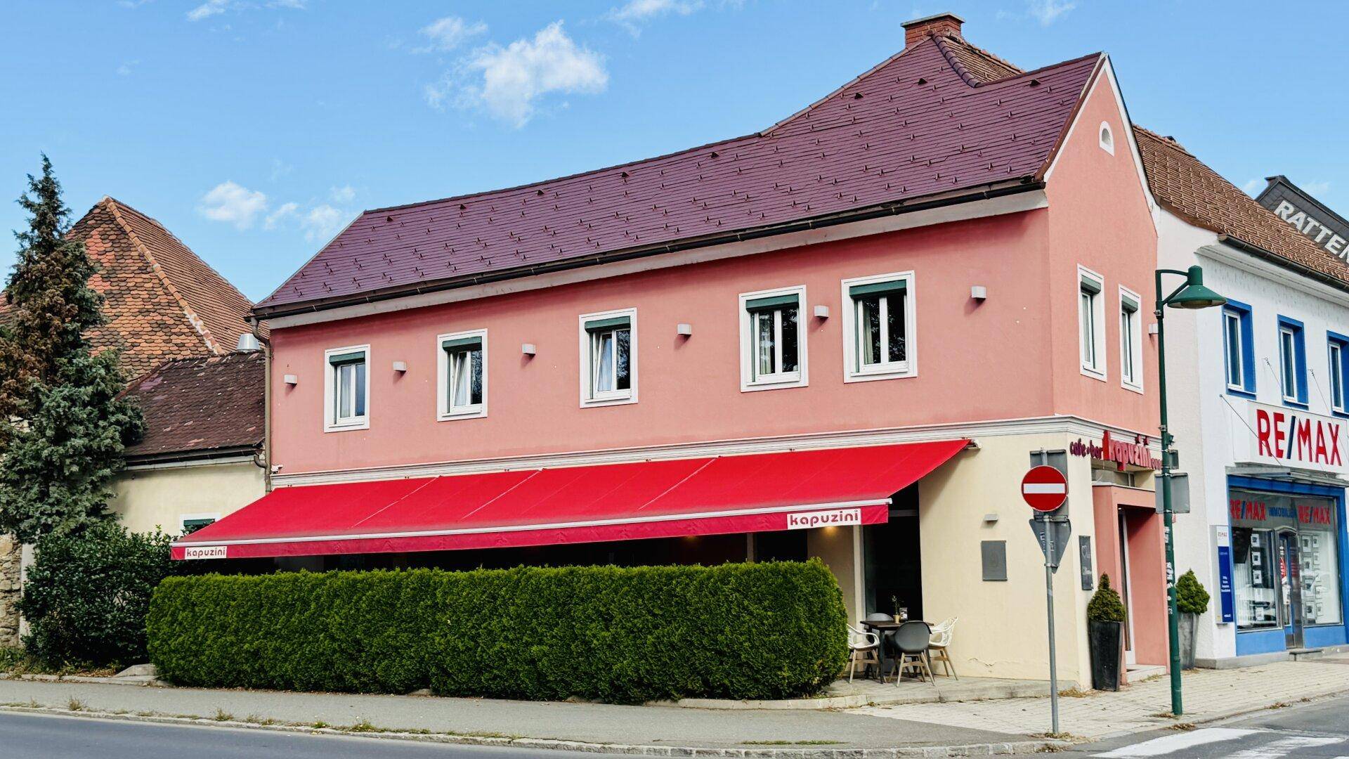 Rosa gestrichenes Gebäude mit roter Markise und gepflegter Hecke vor dem Café.