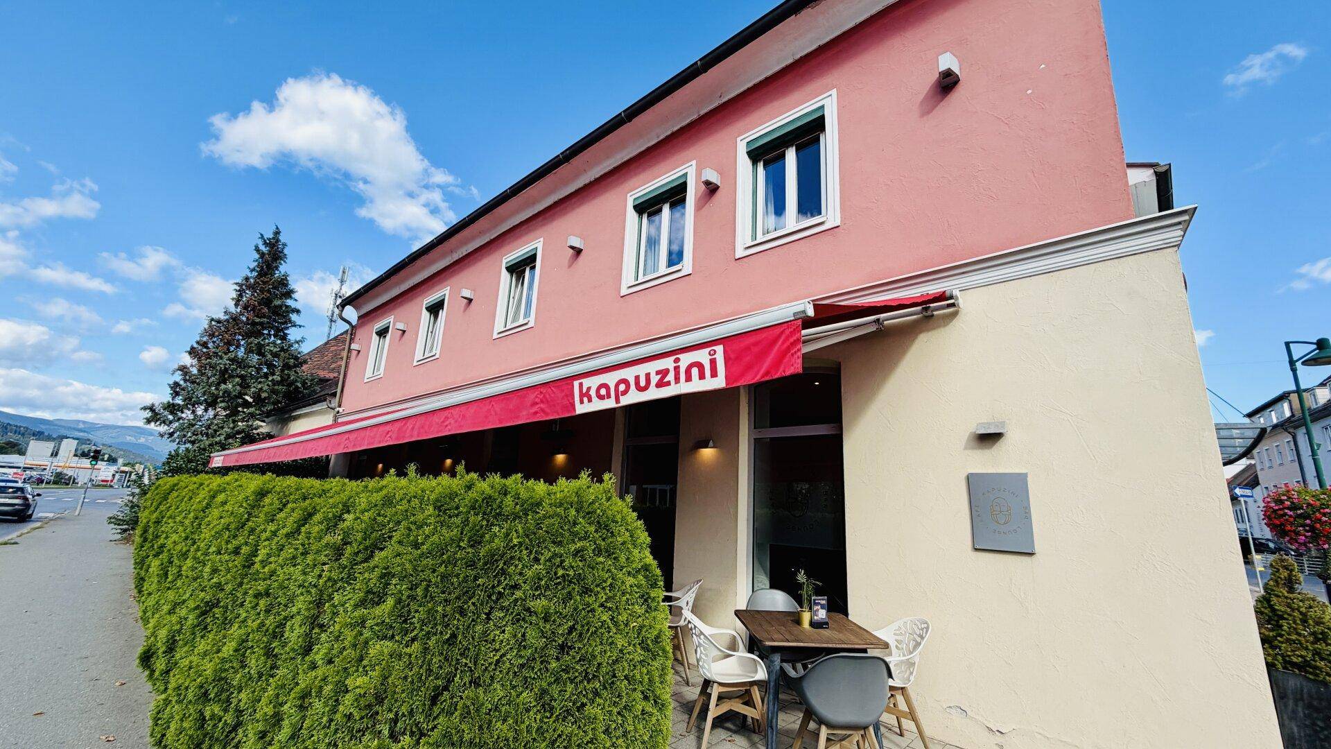 Eingangsbereich des Cafés mit Außenterrasse, Hecke und Blick auf die Straße.
