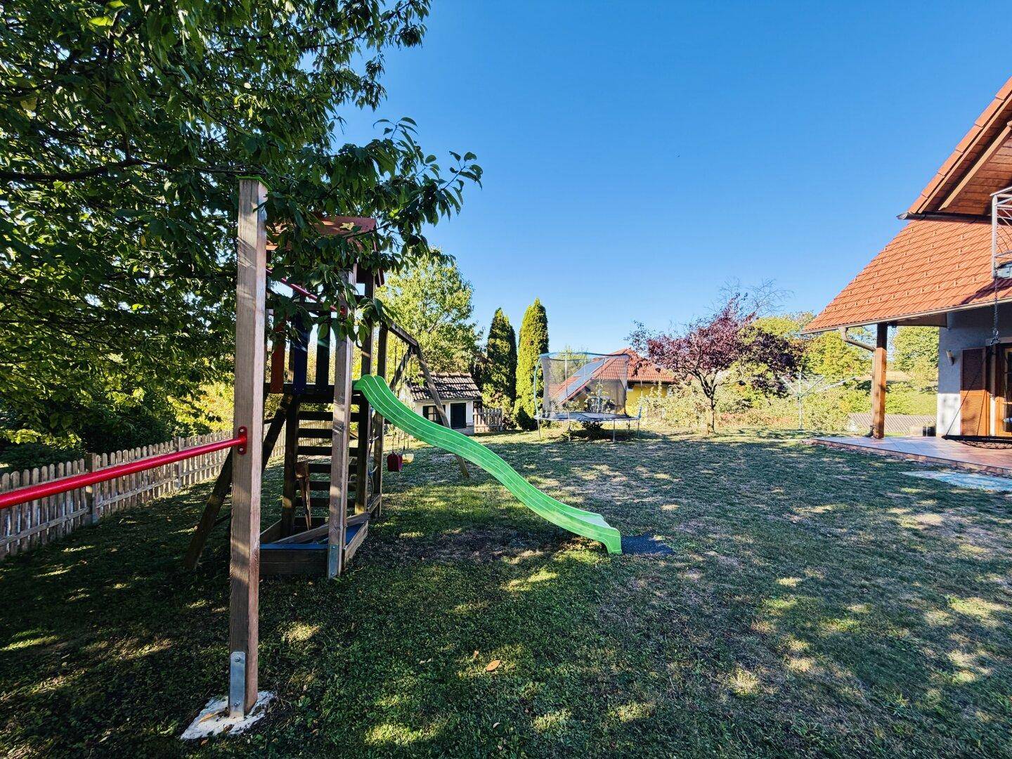 Großer Gartenbereich mit Kinderspielplatz, Rutsche und Schaukel, sowie einem Trampolin im Hintergrund.
