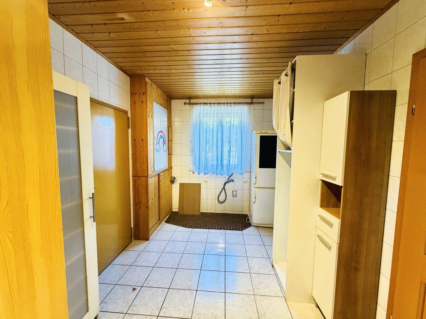 Hauswirtschaftsraum mit Fenster, Fliesenboden und Holzdecke, bereit für Waschmaschinenanschluss.