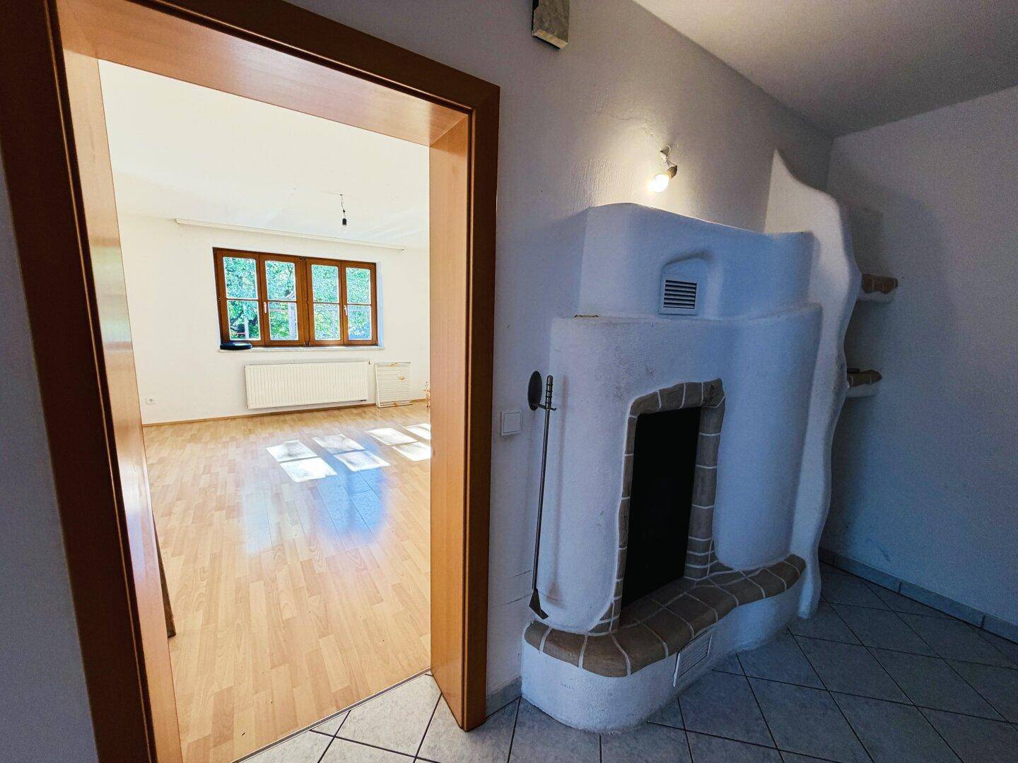 Blick vom Flur in ein helles Wohnzimmer mit Holzboden und einem dekorativen Kachelofen.