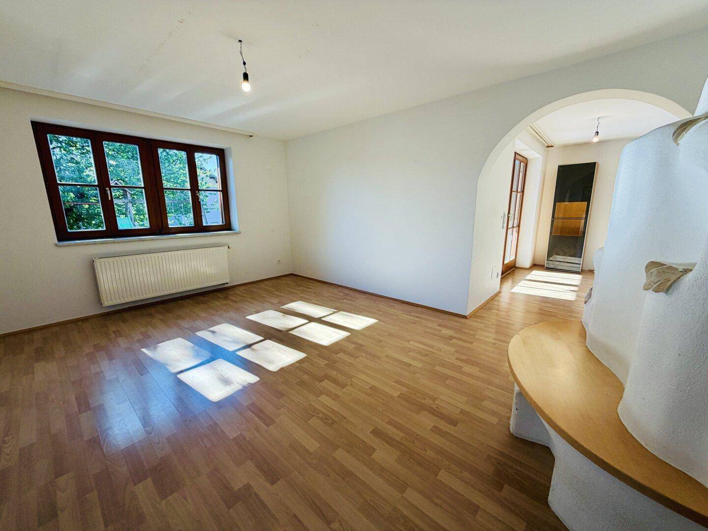 Lichtdurchflutetes Wohnzimmer mit Holzboden, großem Fenster und einem gemütlichen Kachelofen.