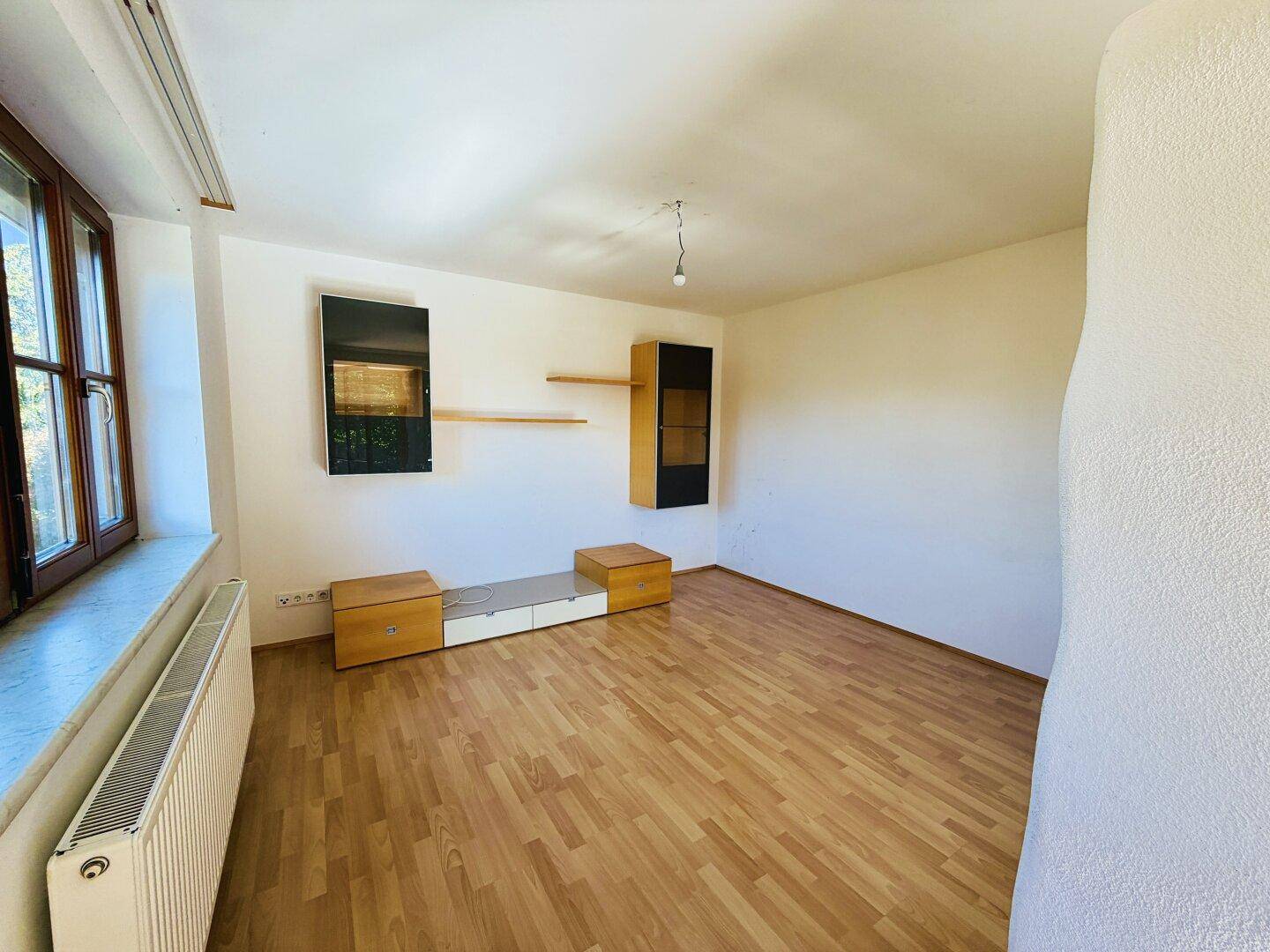 Leeres Wohnzimmer mit Laminatboden, großem Fenster und Heizkörper, bereit zur individuellen Gestaltung.