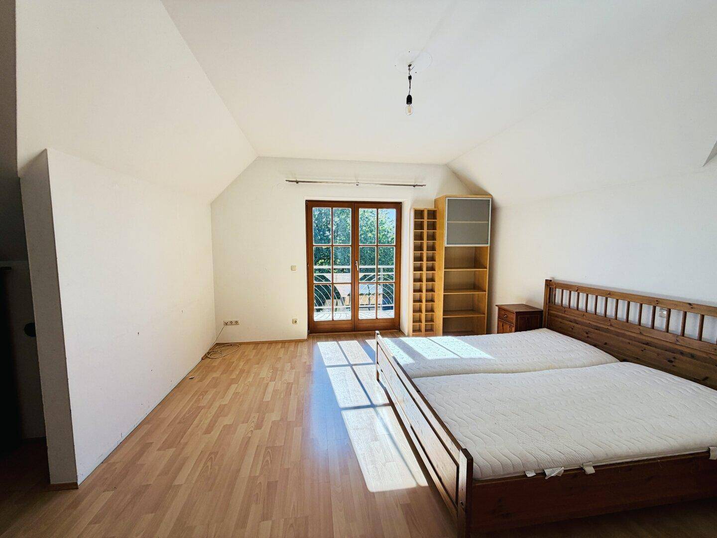 Schlafzimmer mit Holzbett, Laminatboden und Zugang zu einem sonnigen Balkon mit grüner Aussicht.