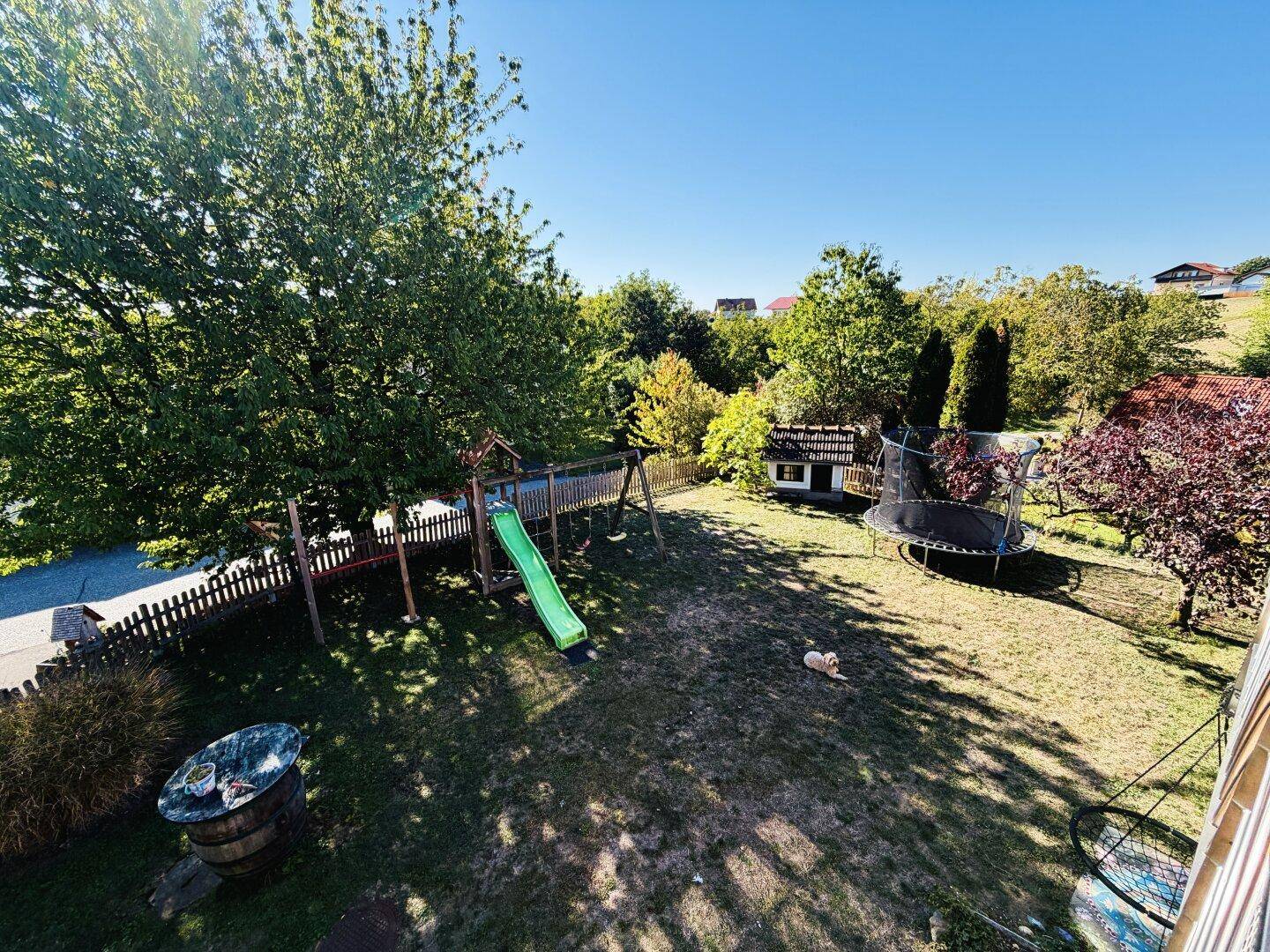 Großer Garten mit Spielplatz, Rutsche und Trampolin, umgeben von Bäumen und viel Grün.