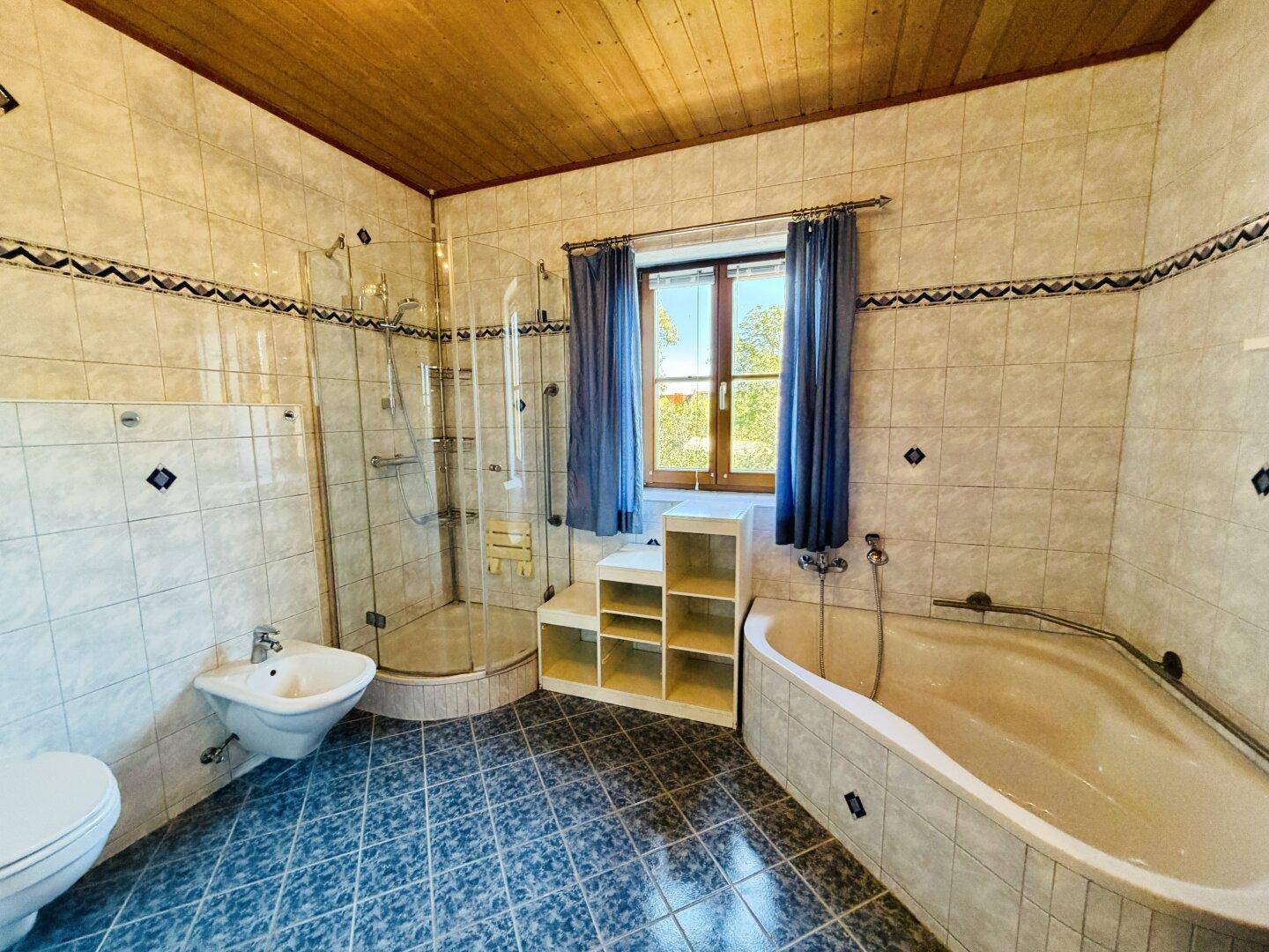 Geräumiges Badezimmer mit Eckbadewanne, Duschkabine, Bidet und Fenster, gefliest in hellen und blauen Tönen.