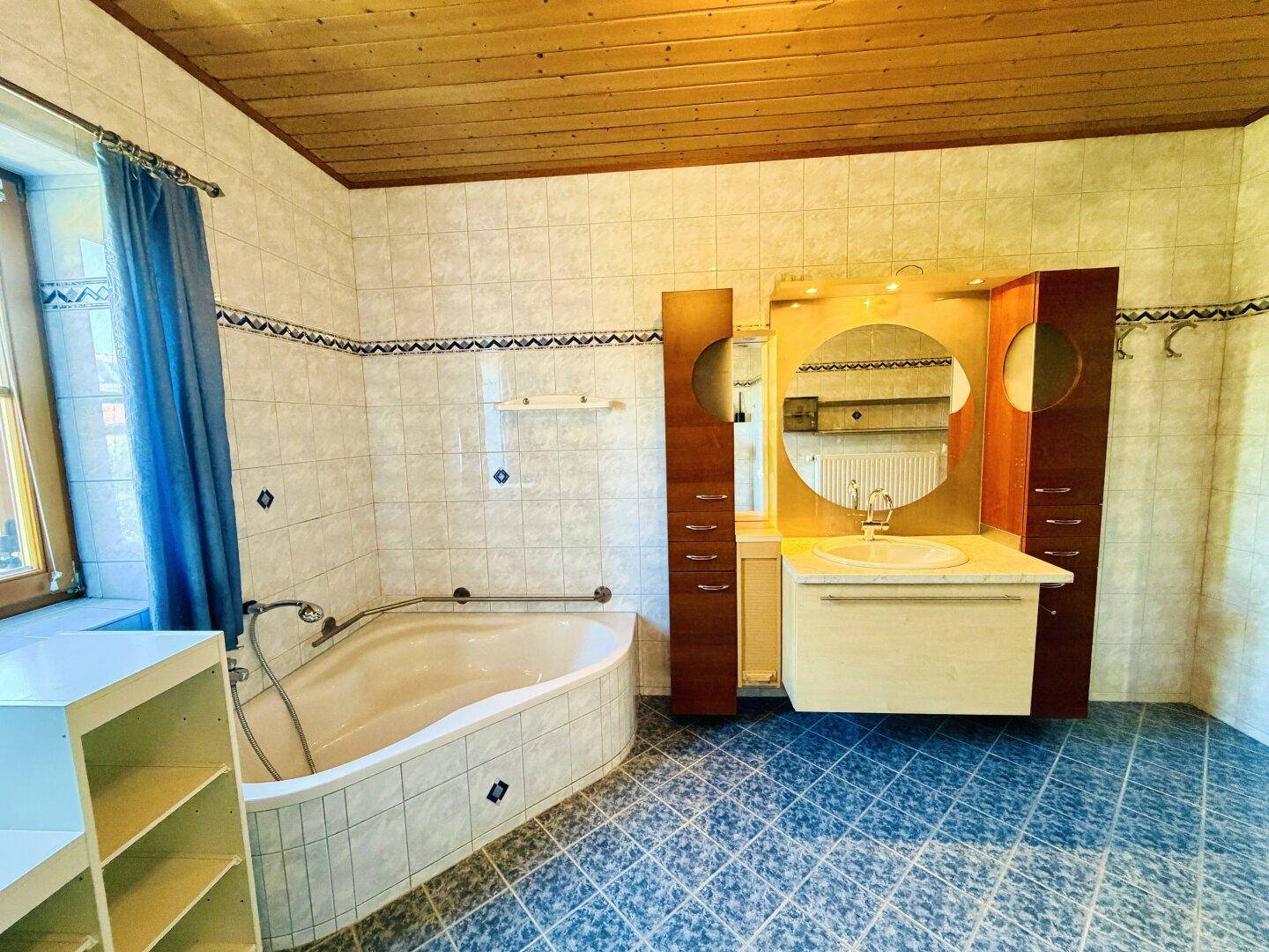 Badezimmer mit Eckbadewanne, Waschtisch mit Spiegel und Holzdecke, gefliest in hellen und blauen Tönen.