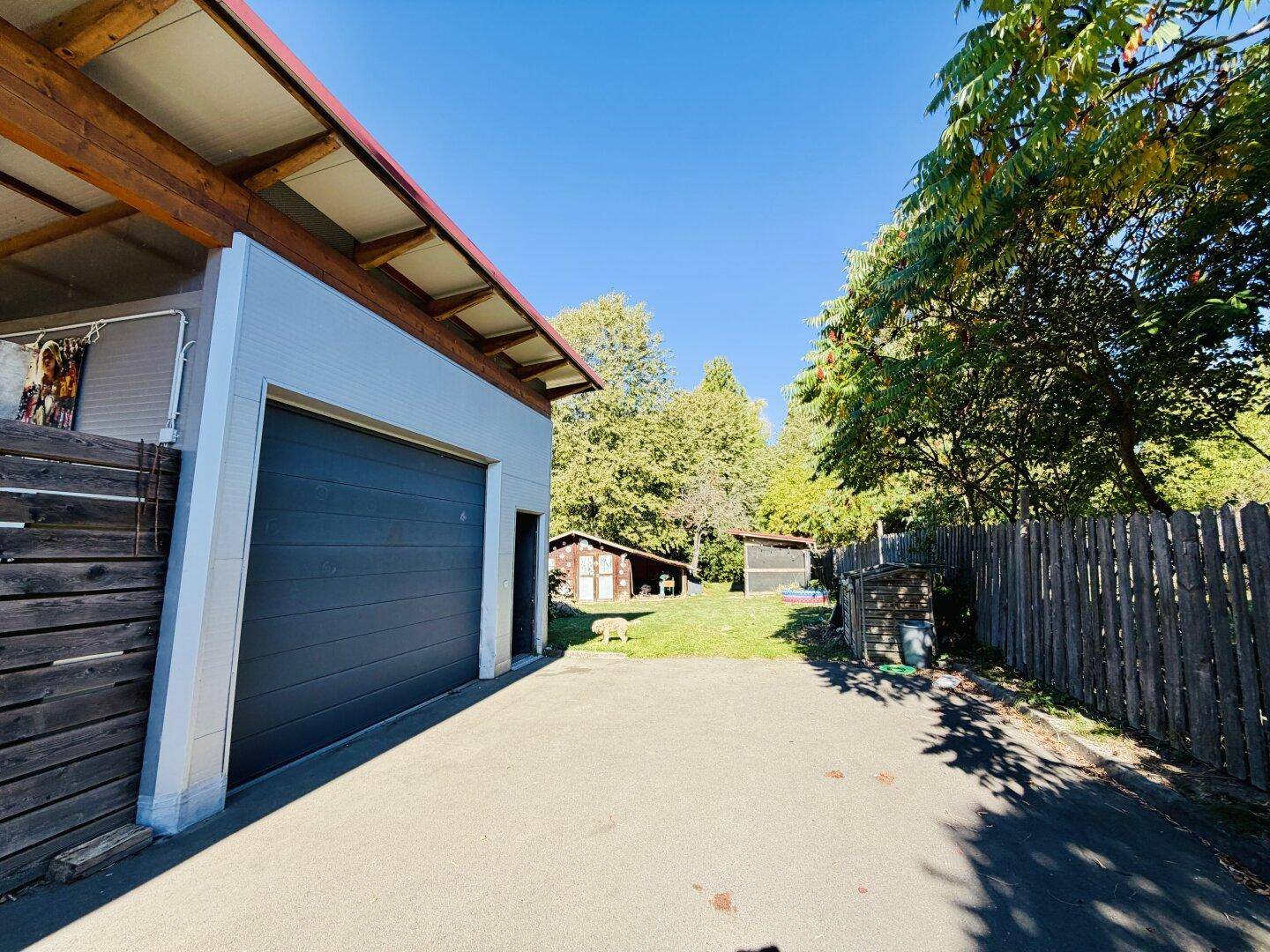 Garage mit dunklem Tor und Blick auf den Hinterhof mit Holzschuppen und Bäumen.