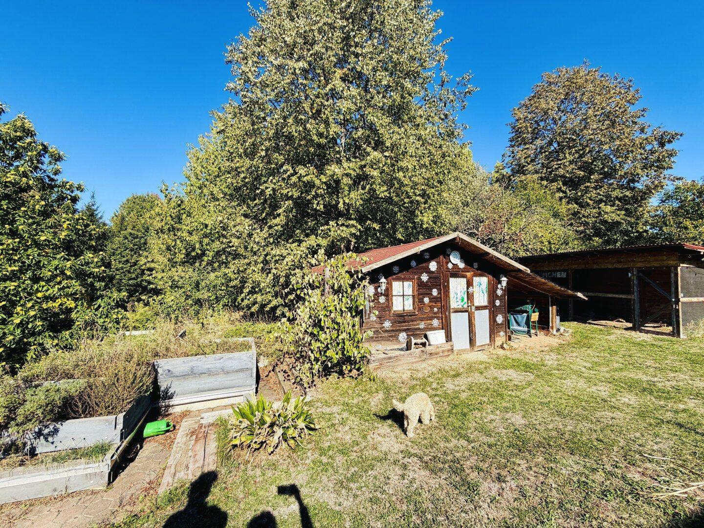 Gartenbereich mit Hochbeeten, einem kleinen Holzspielhaus und alten Bäumen im Hintergrund.