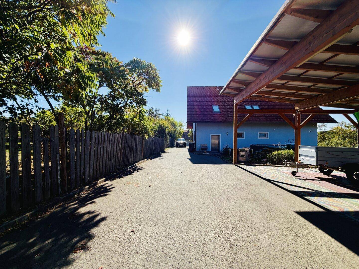 Zufahrt zum Haus und Carport, begrenzt durch einen Holzzaun, unter blauem Himmel.