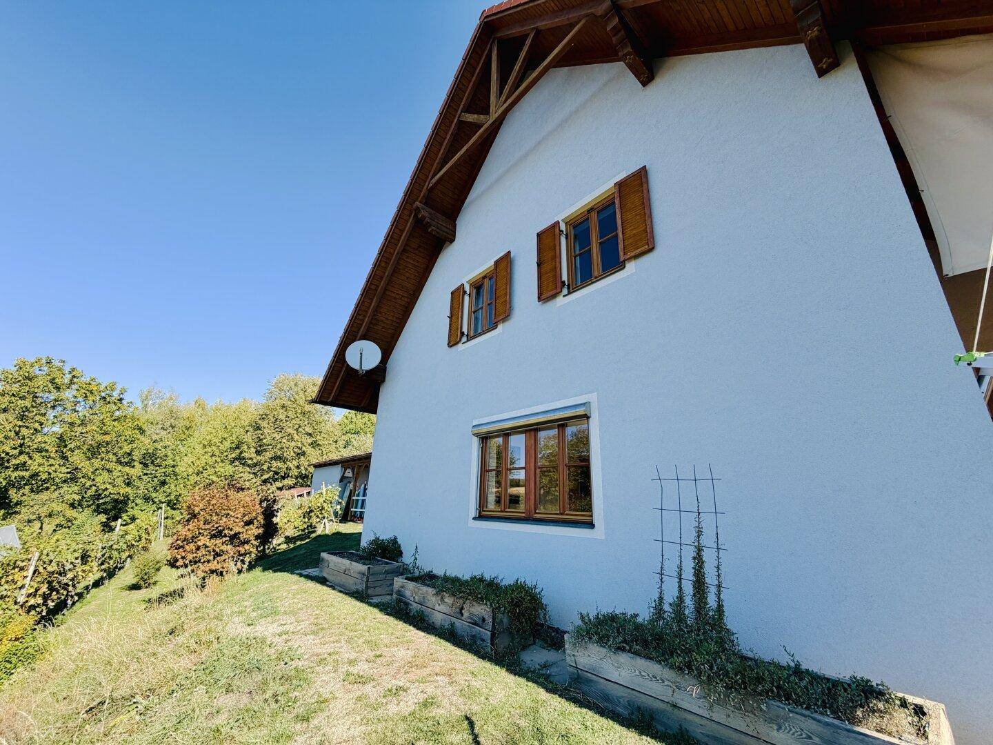 Seitenansicht des Hauses mit Holzfenstern, Fensterläden und terrassiertem Gartenbereich.