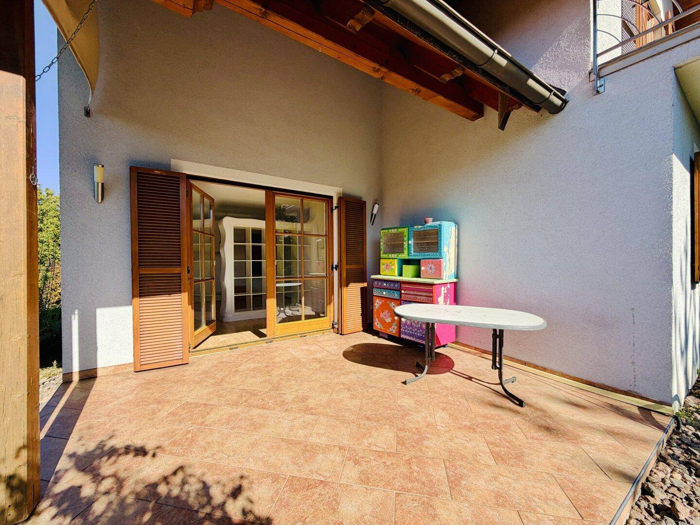 Überdachte Terrasse mit Fliesenboden und geöffneten Doppeltüren zum Innenbereich des Hauses.