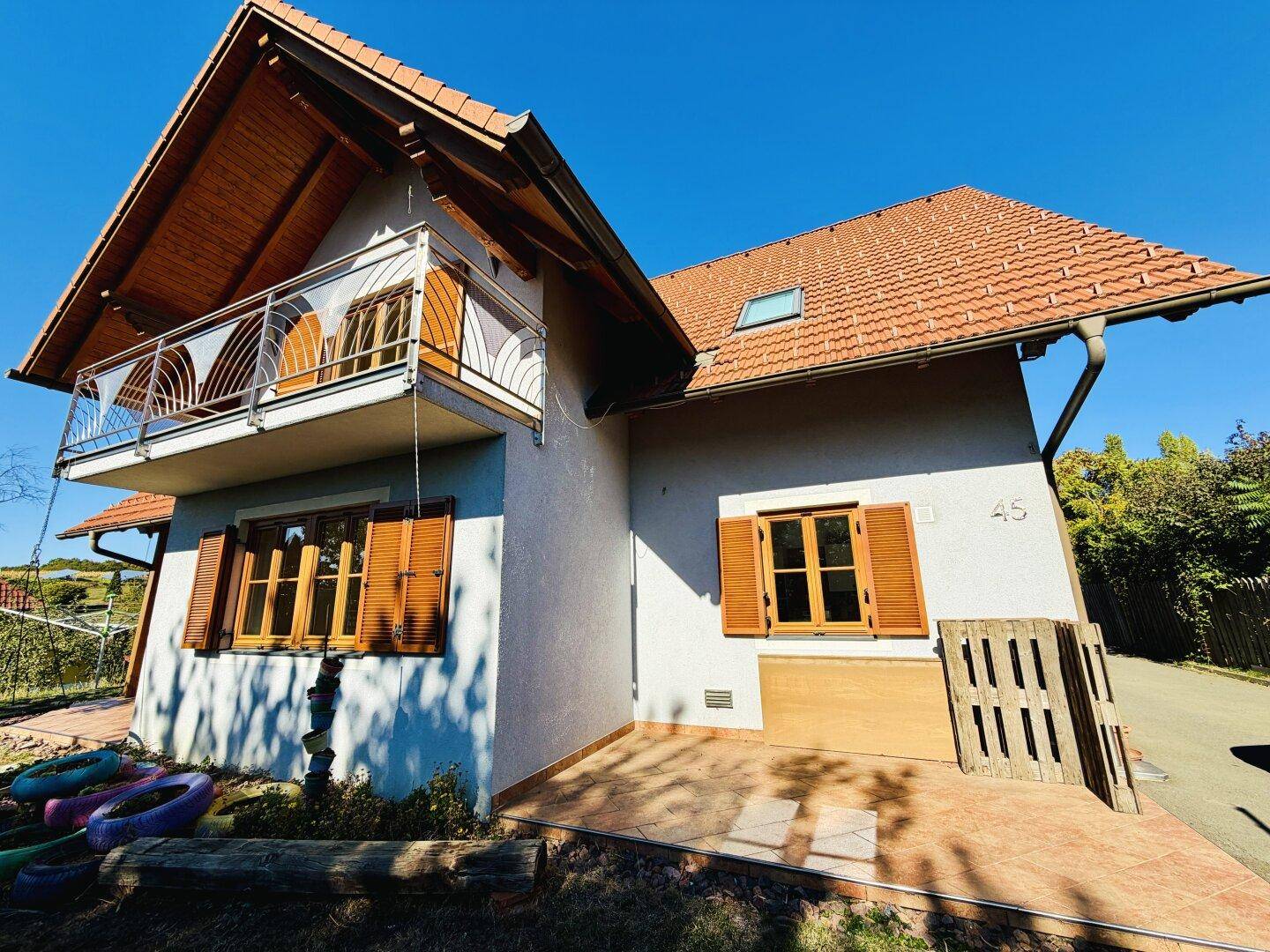 Ansicht des Hauses mit Balkon, Holzfenstern und Fensterläden sowie einem kleinen gepflasterten Bereich.