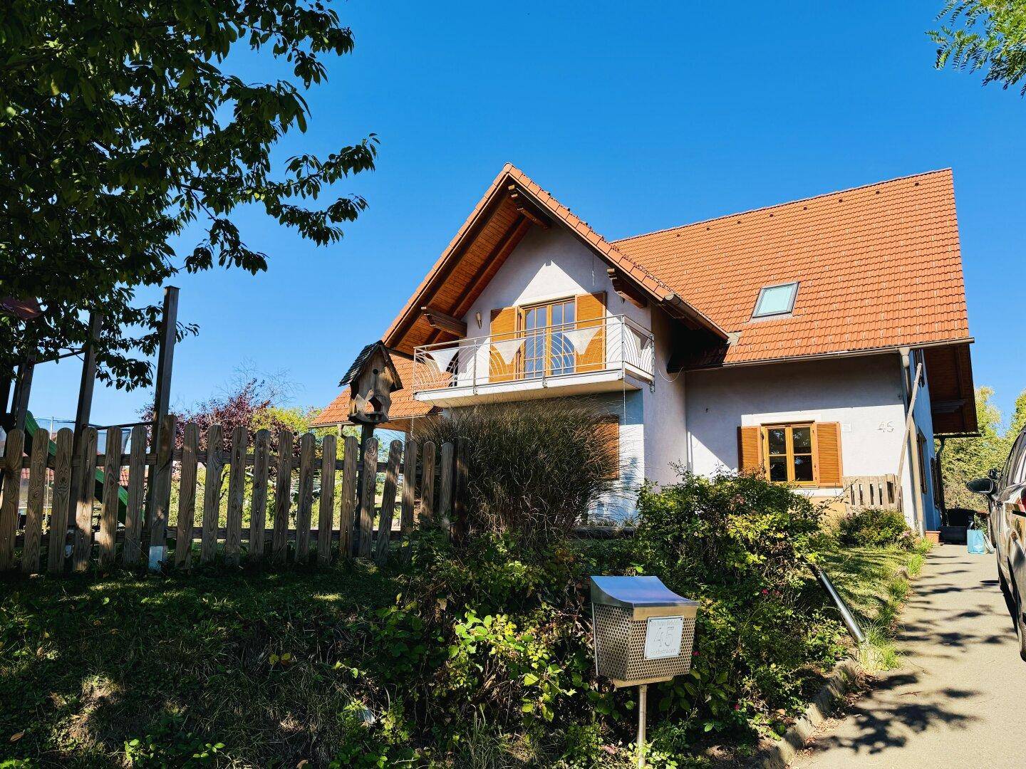 Vorderansicht des Hauses von der Straße aus, umgeben von einem Holzzaun und Büschen.