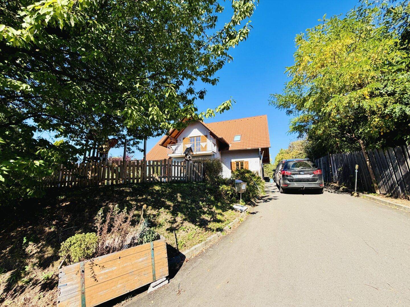 Weitwinkelansicht des Hauses von der Zufahrt aus, mit geparktem Auto und viel Grün.