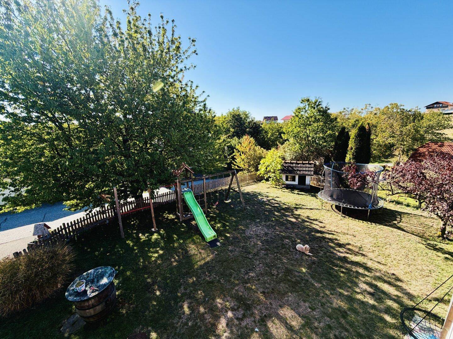 Großer Gartenbereich mit Kinderspielplatz, Rutsche, Schaukeln und einem Trampolin unter blauem Himmel.