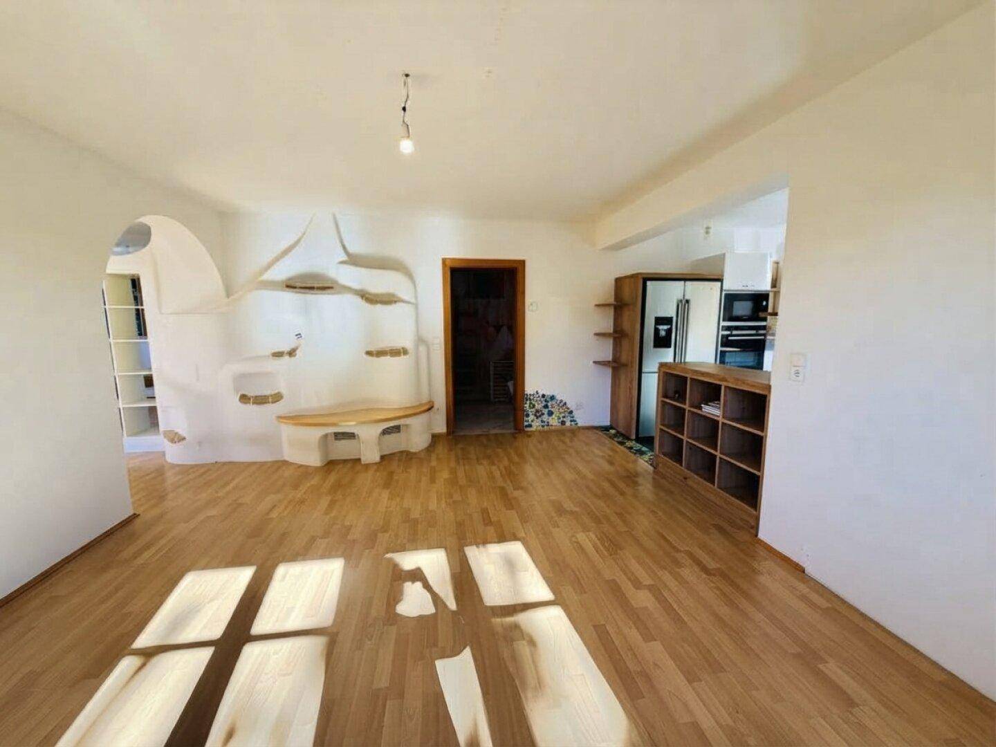 Geräumiges Wohnzimmer mit Holzboden, Kachelofen und Blick auf den offenen Küchenbereich.