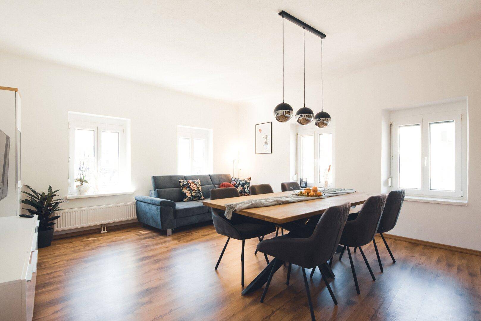 Heller Wohn- und Essbereich mit Holzboden, Sofa und großem Esstisch unter Hängelampen.