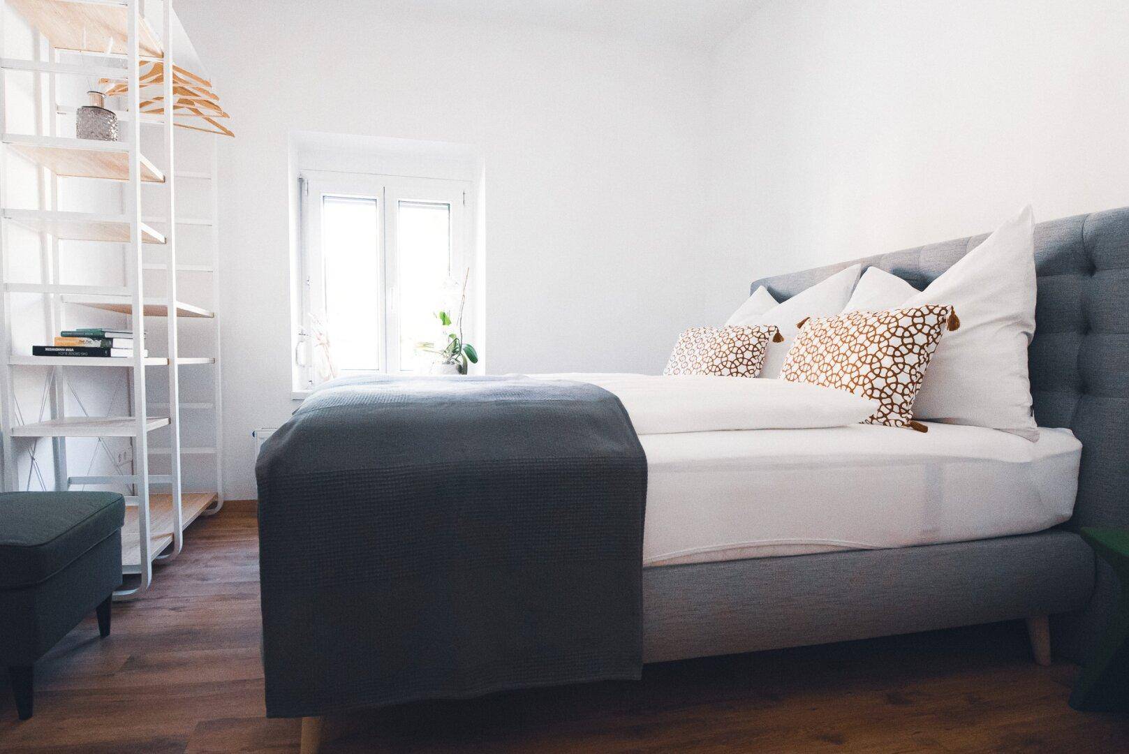 Helles Schlafzimmer mit Doppelbett, Holzboden und offenem Regalsystem am Fenster.