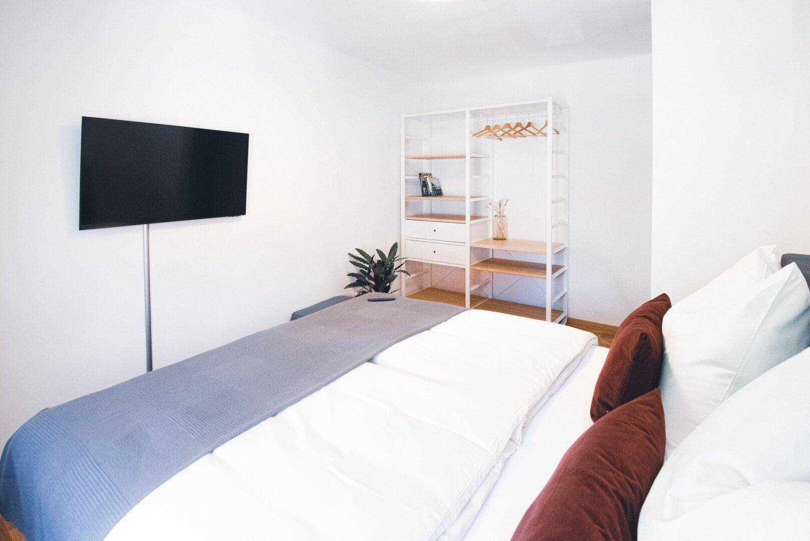 Schlafzimmer mit Doppelbett, Wandfernseher und offenem Kleiderschrank auf Holzboden.