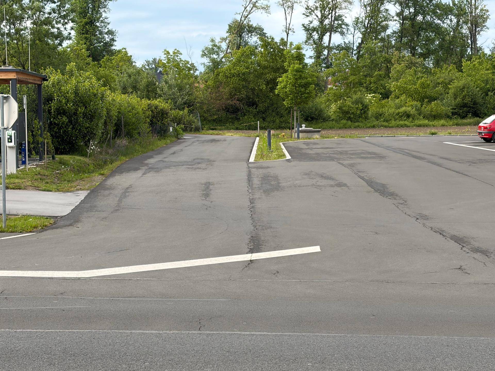 Einfahrt zum Grundstück mit asphaltiertem Bereich, der zu einem Parkplatz führt, umgeben von Bäumen und Büschen.