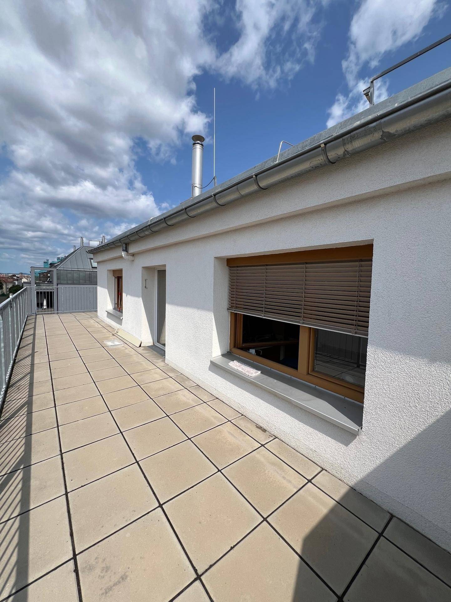 Sonnige Dachterrasse mit Fliesenboden und Fenstern mit Außenjalousien, die einen hellen Außenbereich bieten.