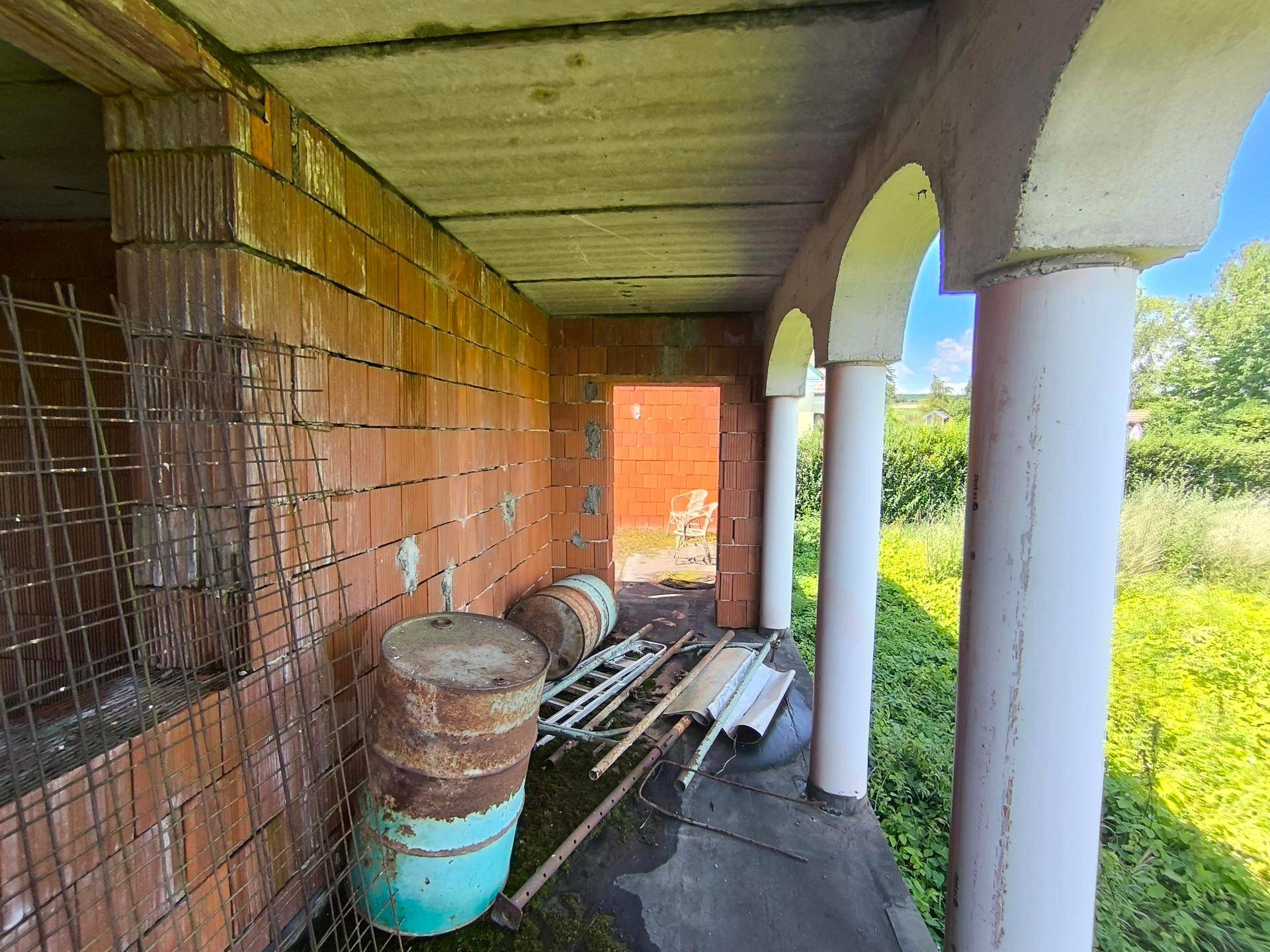 Überdachter Bereich im Rohbau mit Säulen und Blick auf die verwilderte Außenanlage.