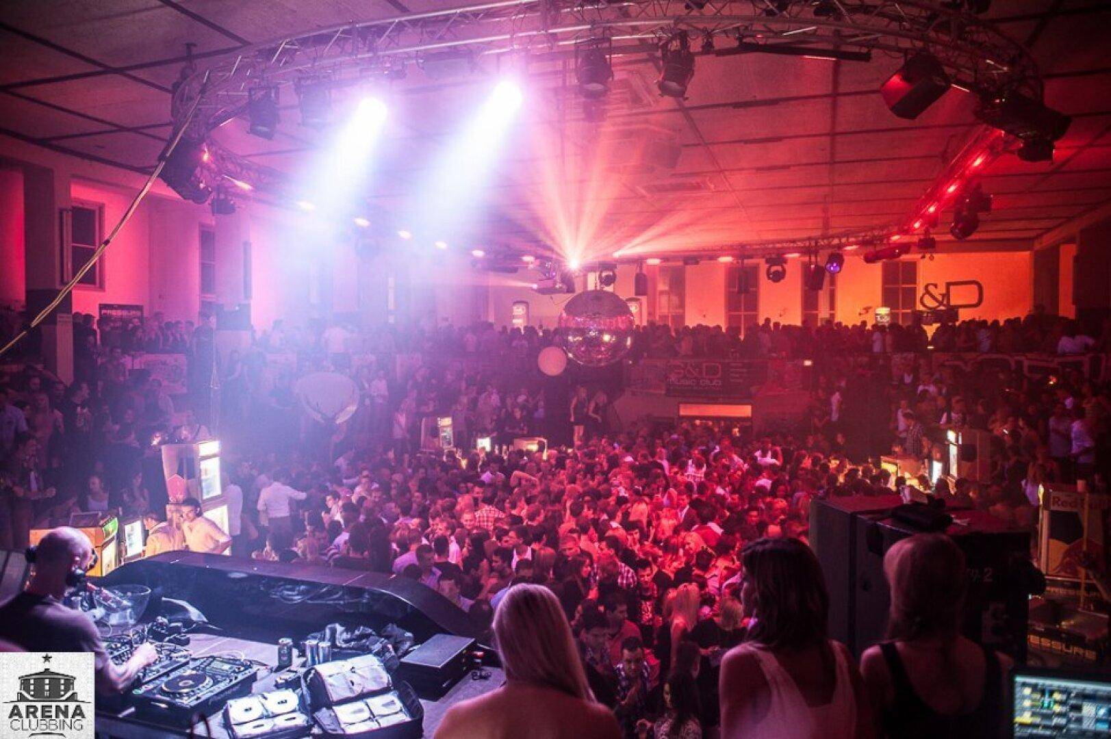 Großer Clubraum mit vielen tanzenden Menschen, DJ-Pult und beeindruckender Lichtshow, die den Raum erhellt.