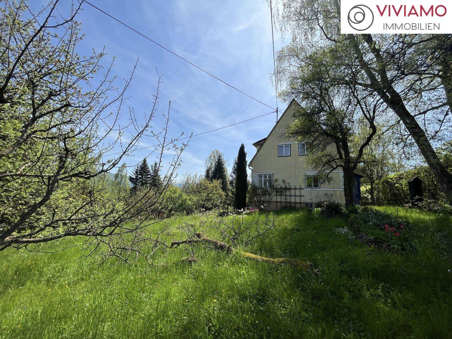 Freistehendes Haus mit Satteldach, umgeben von einem großen grünen Garten und Bäumen unter blauem Himmel.