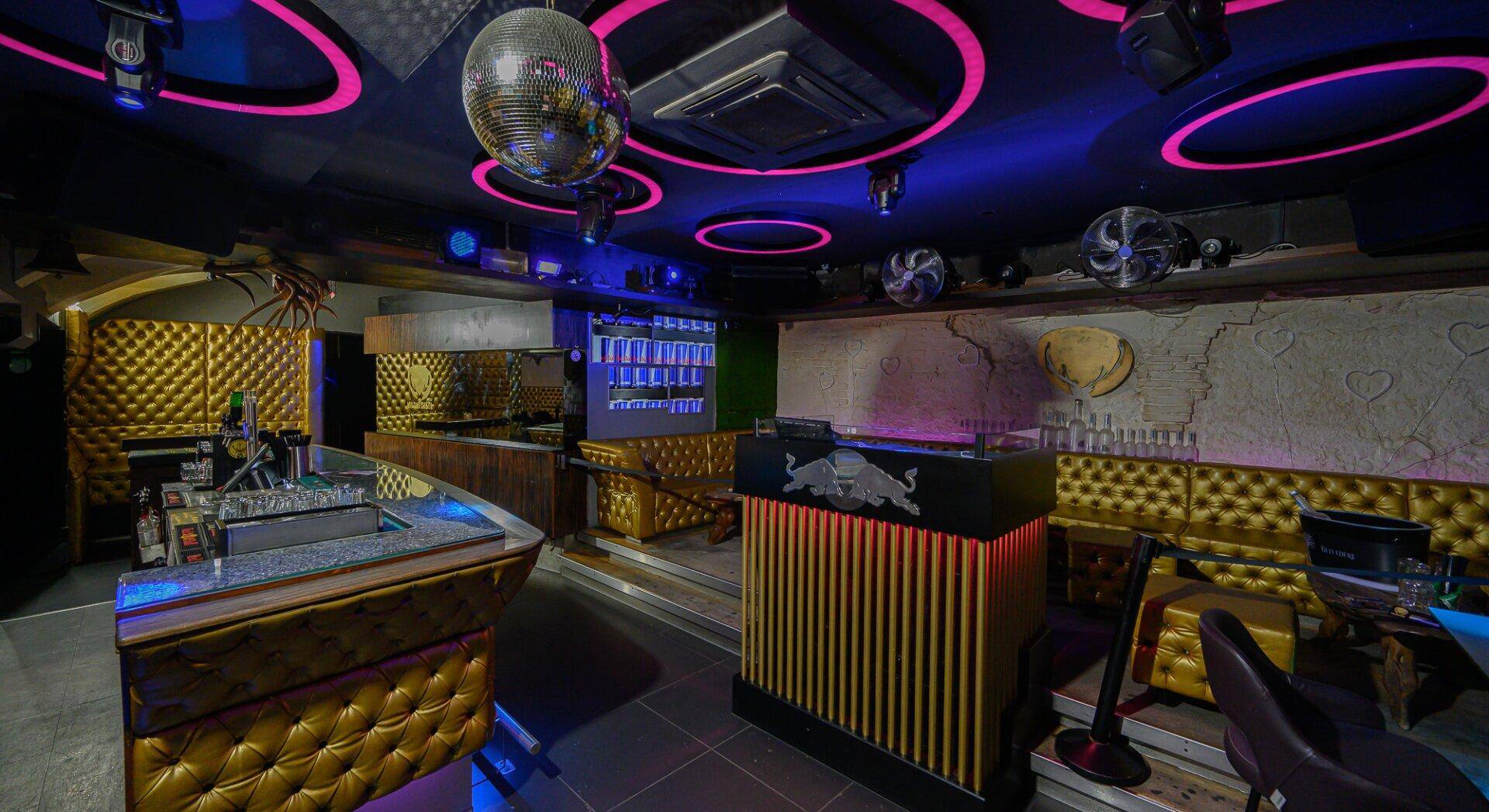 Innenansicht einer Bar mit goldgepolsterten Sitzbereichen, DJ-Pult und Deckenbeleuchtung, die eine dynamische Atmosphäre schafft.