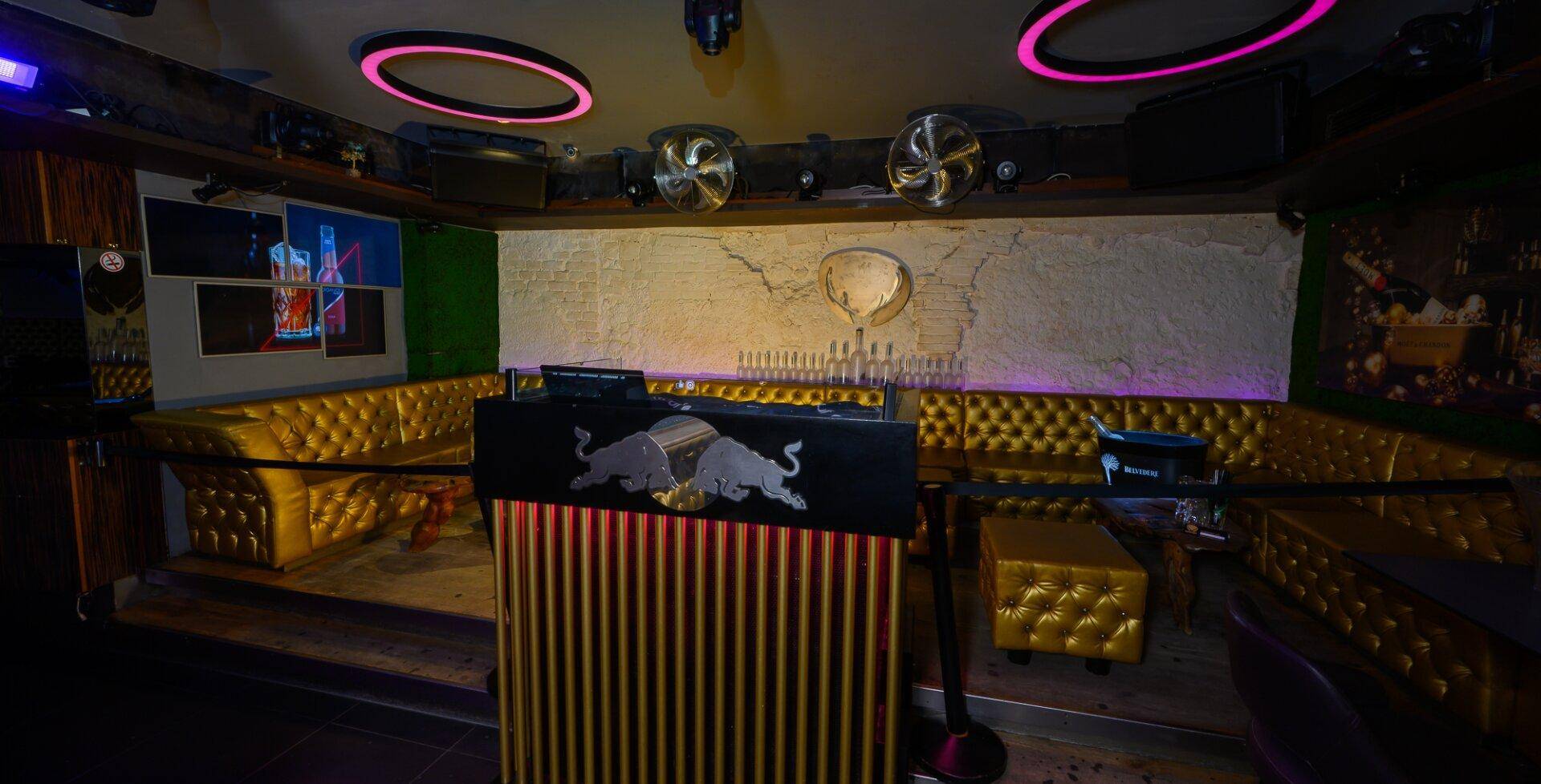Sitzbereich einer Bar mit goldgepolsterten Bänken, DJ-Pult und Deckenbeleuchtung, die eine lebhafte Atmosphäre schafft.