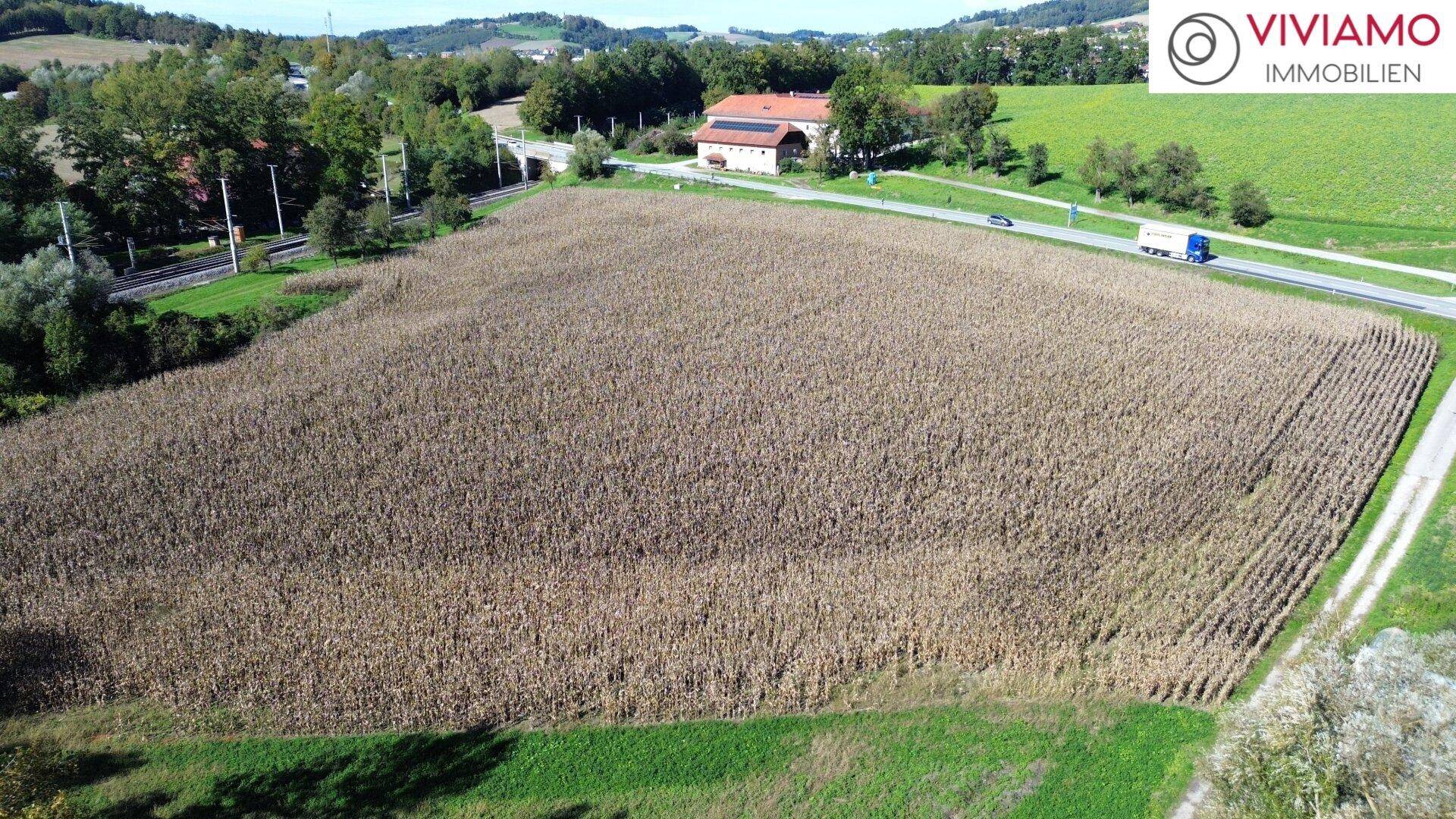 Weitläufige Luftaufnahme eines Feldes, umgeben von ländlichen Straßen und Bahngleisen, mit Gebäuden in der Ferne.