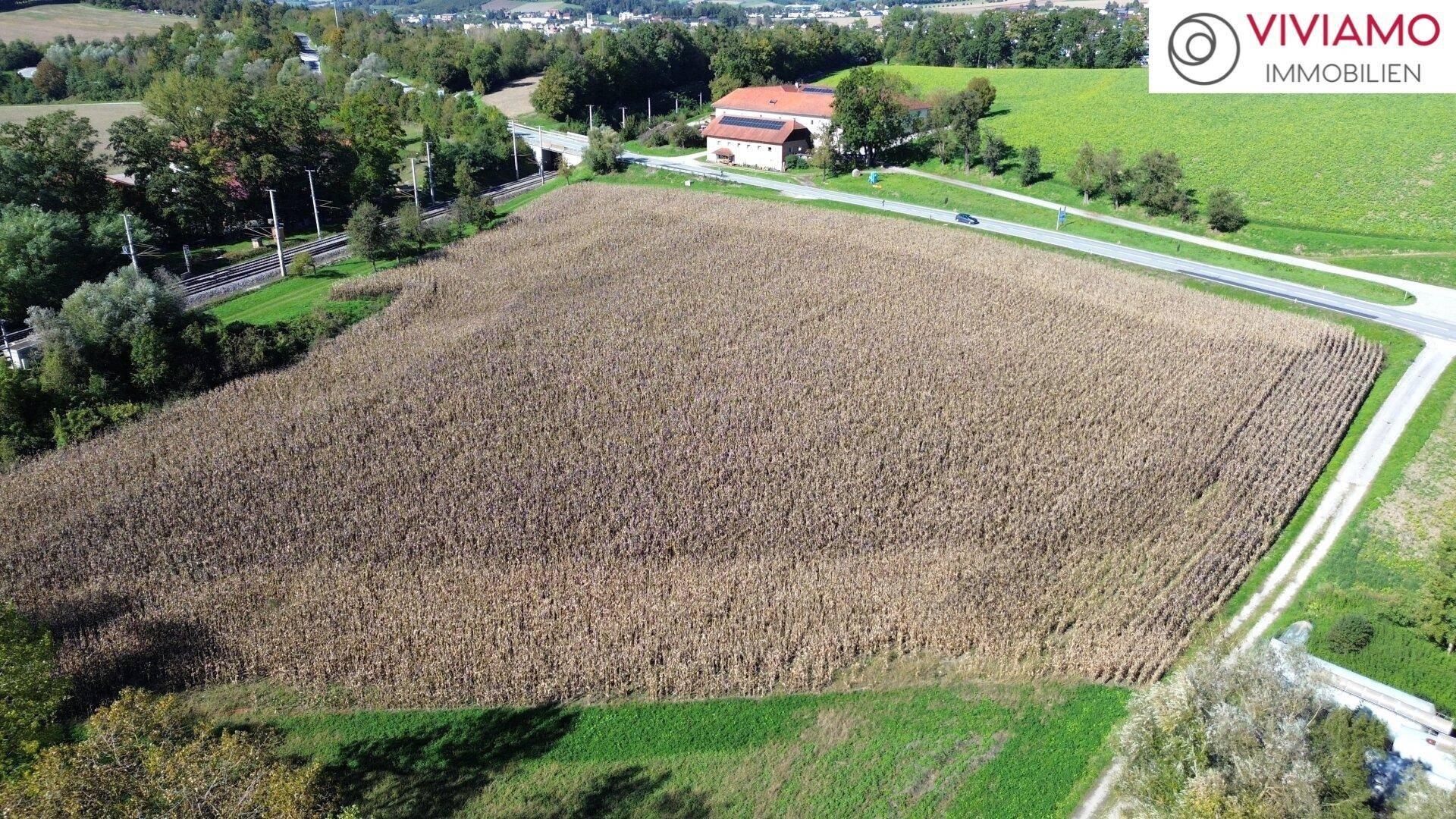 Luftperspektive auf ein großes Feld, angrenzende Straßen und Bahngleise, mit ländlicher Bebauung im Hintergrund.