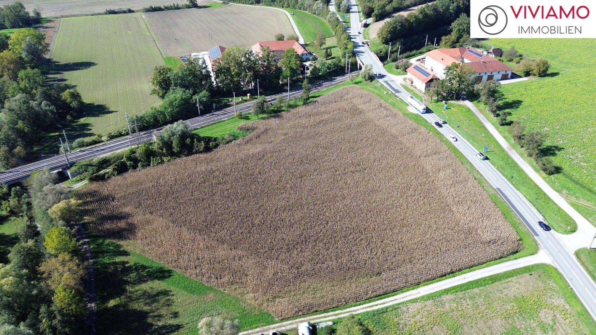 Übersicht aus der Vogelperspektive auf ein großes Feld, angrenzende Straßen, Bahngleise und ländliche Bebauung.