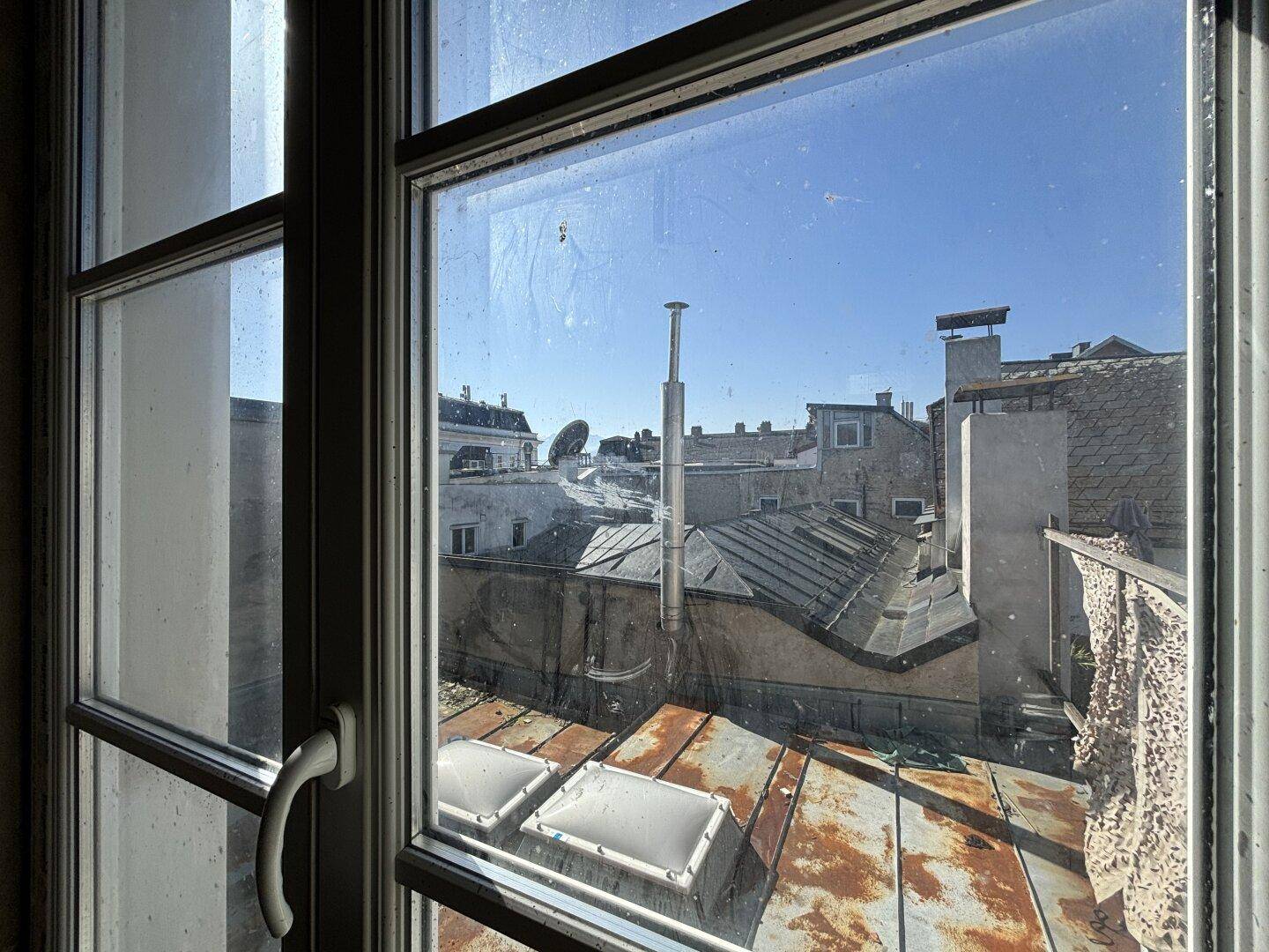 Blick aus einem Fenster über die Dächer der Nachbargebäude unter strahlend blauem Himmel.