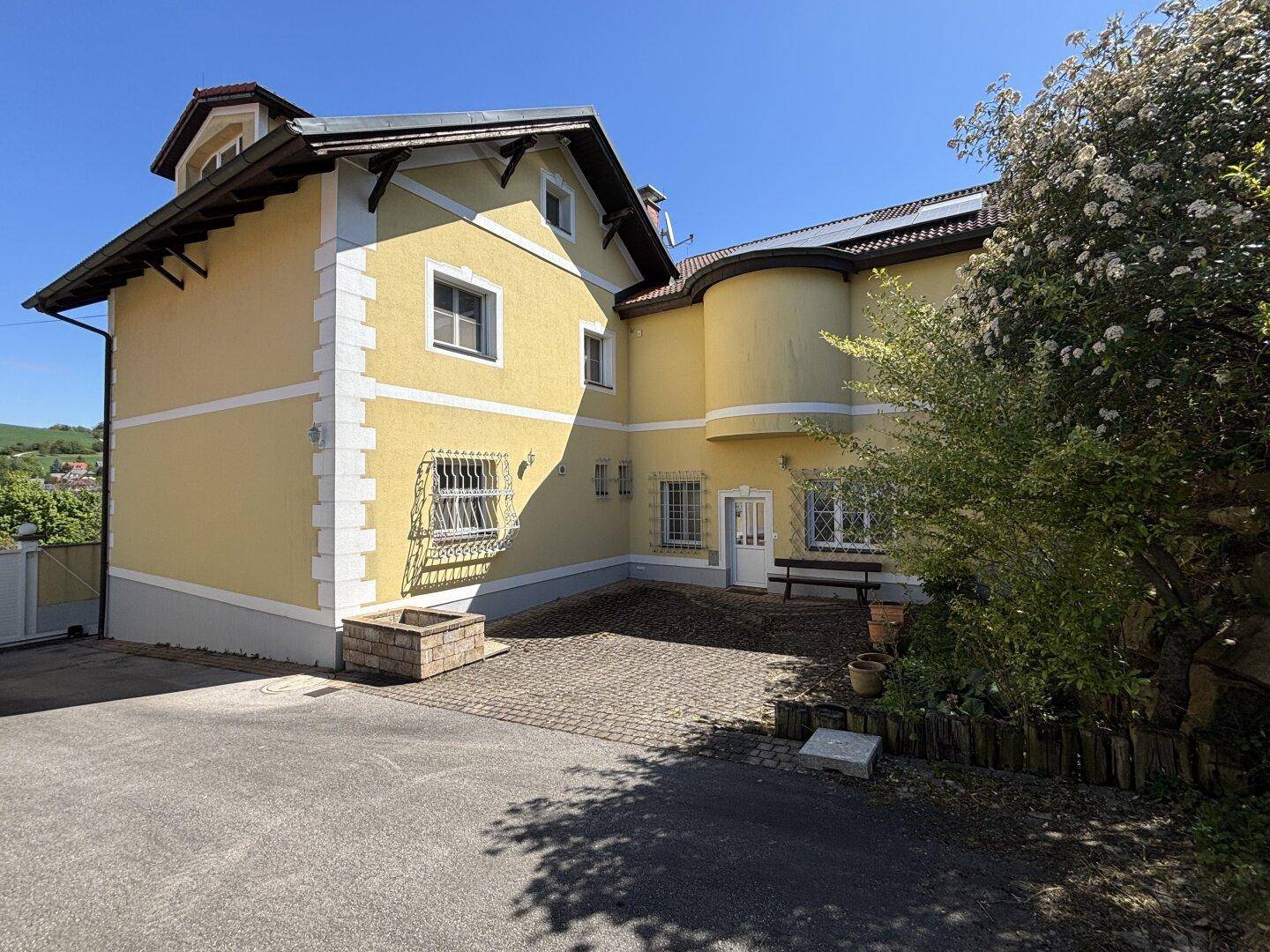 Seitenansicht der gelben Villa mit gepflastertem Bereich und Blick auf die Umgebung.