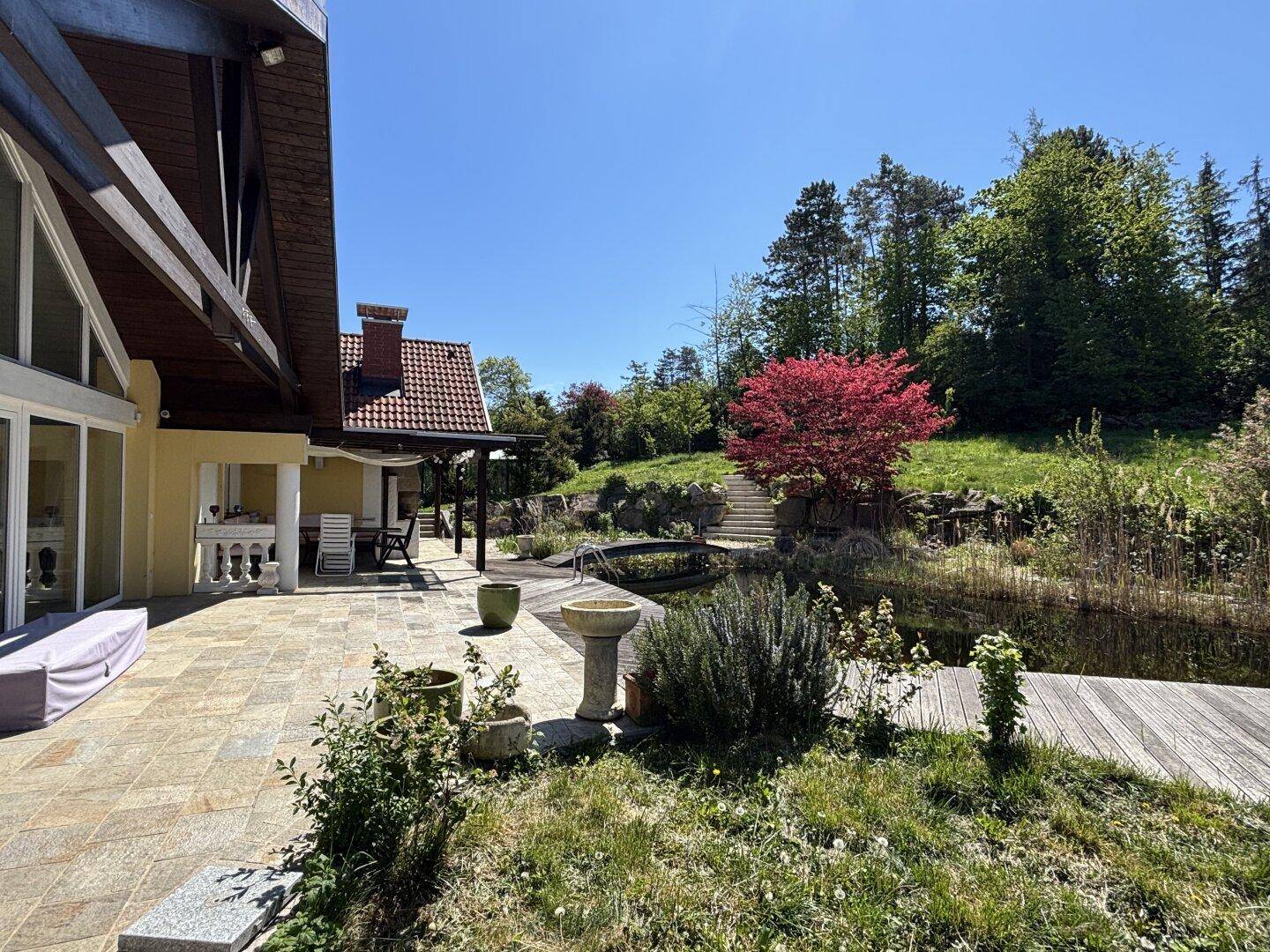 Große Steinterrasse mit Sitzgelegenheiten und Blick auf den Garten mit Teich und Bäumen.