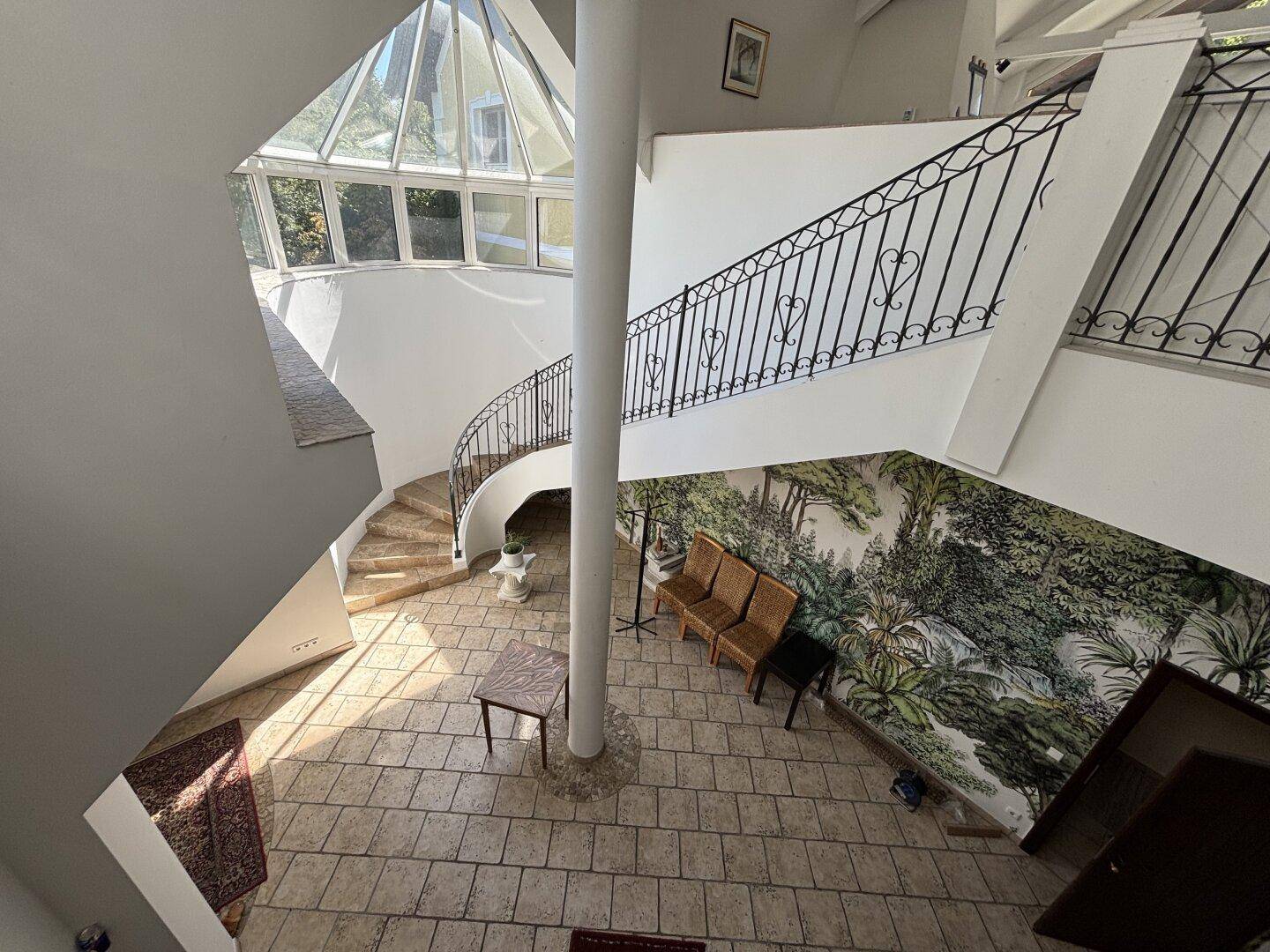 Heller Eingangsbereich mit geschwungener Treppe, Glasdach und dekorativer Wandmalerei.