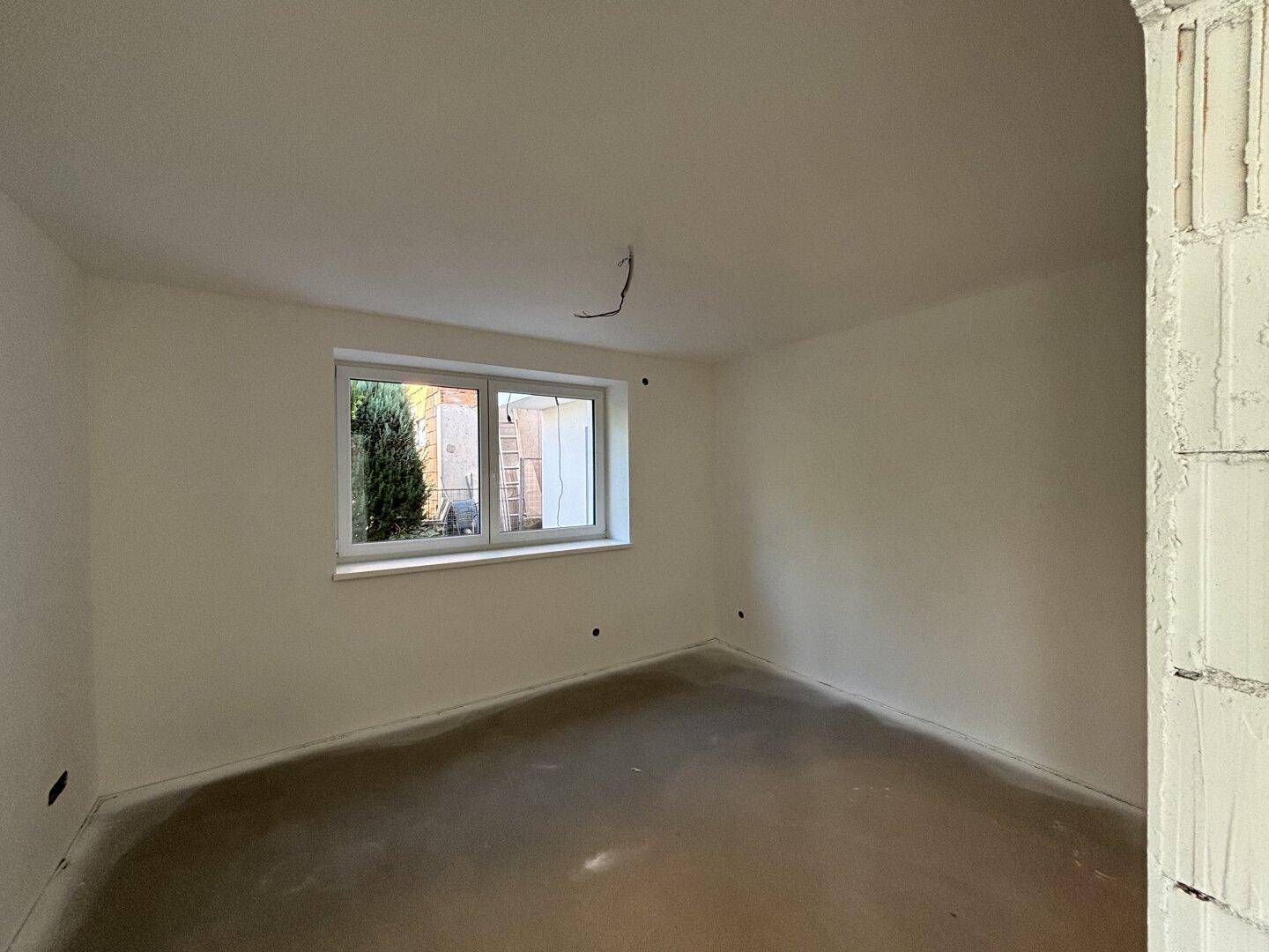 Zimmer im Rohbau mit Fenster und Blick auf die grüne Hecke im Außenbereich.