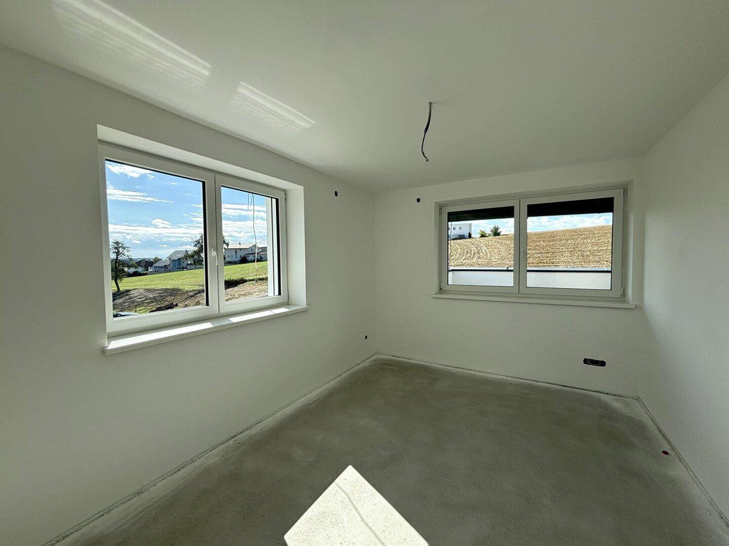 Helles, unfertiges Schlafzimmer mit weißen Wänden, Rohbetonboden und zwei Fenstern mit Ausblick.