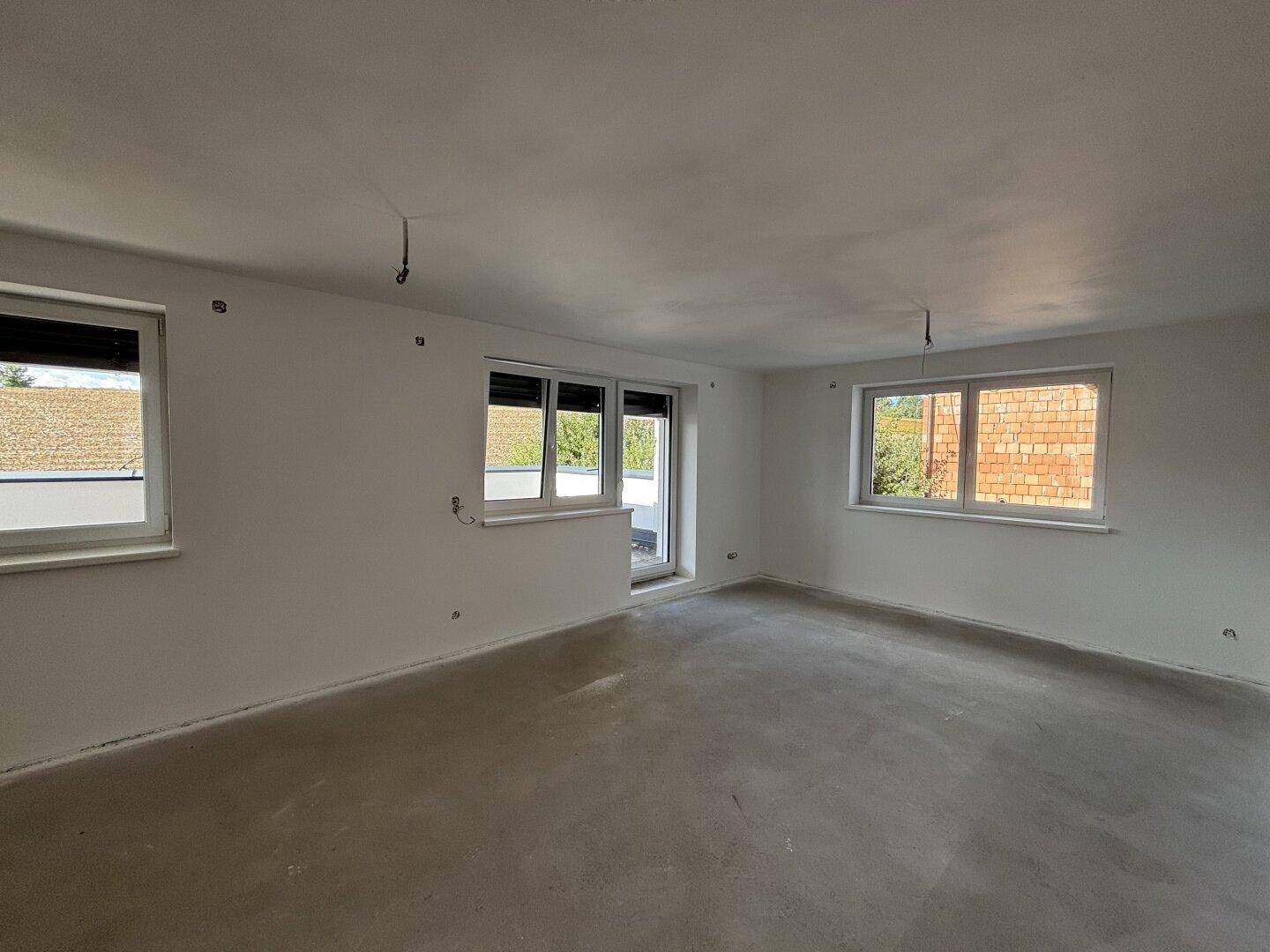 Geräumiges, unfertiges Wohnzimmer mit weißen Wänden, Rohbetonboden und mehreren Fenstern.