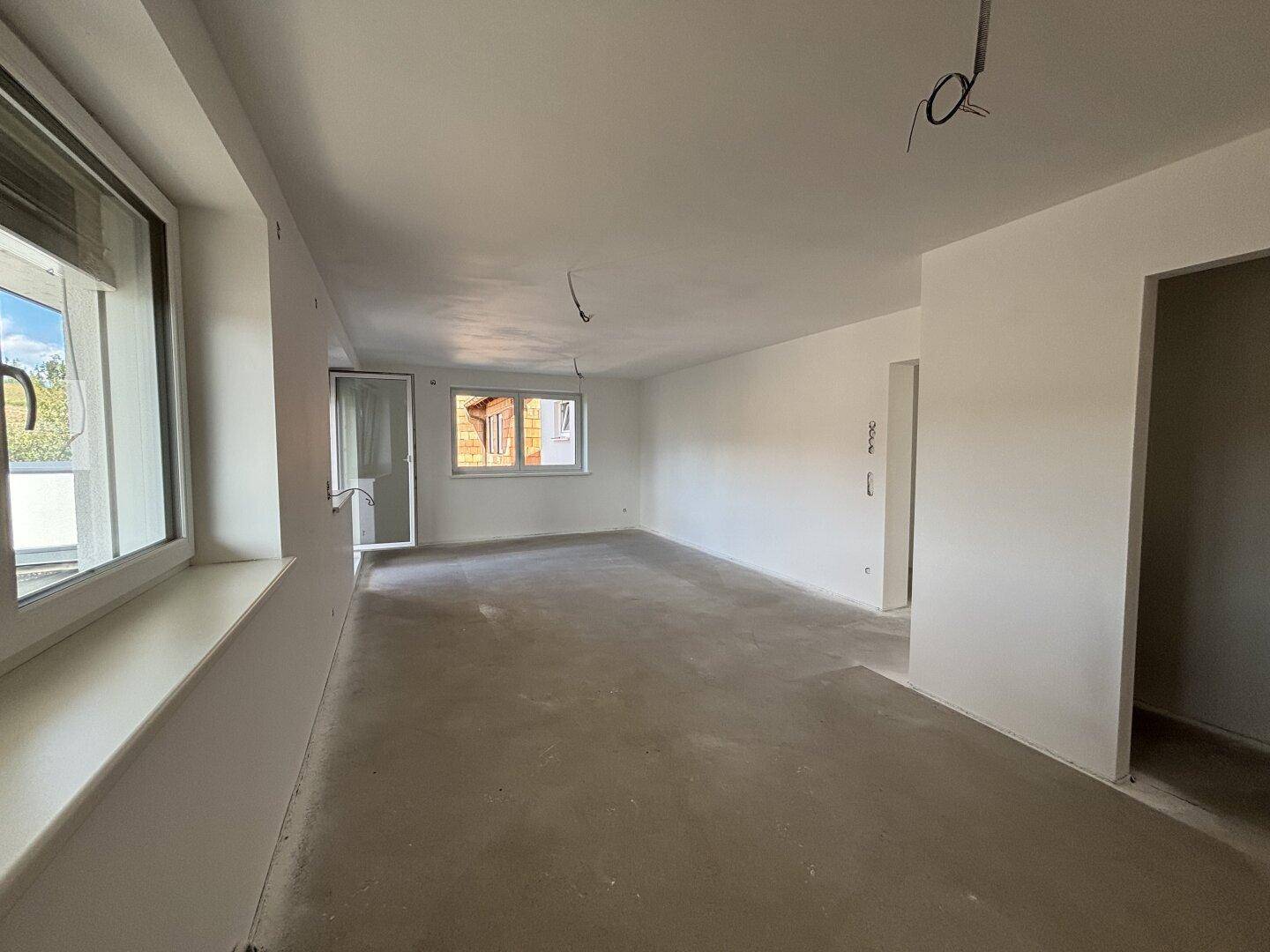 Heller, unfertiger Wohnbereich mit Rohbetonboden und großen Fenstern, die viel Tageslicht hereinlassen.