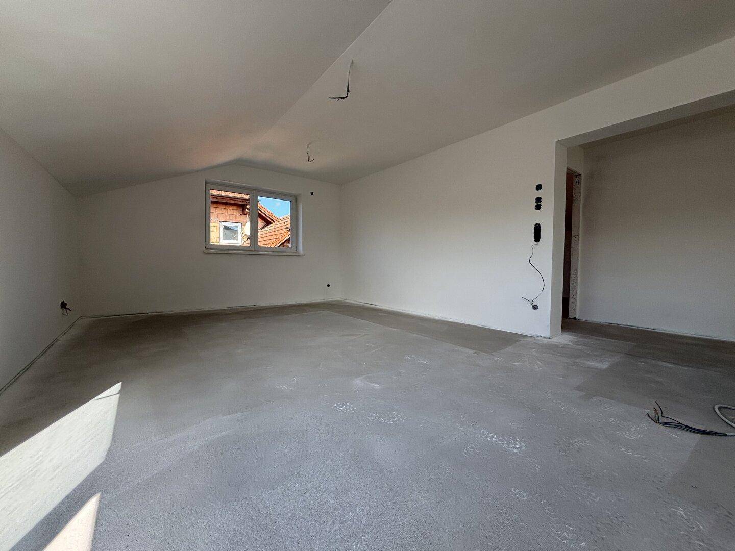 Heller, unfertiger Raum im Dachgeschoss mit Dachschrägen und Blick auf die Umgebung.