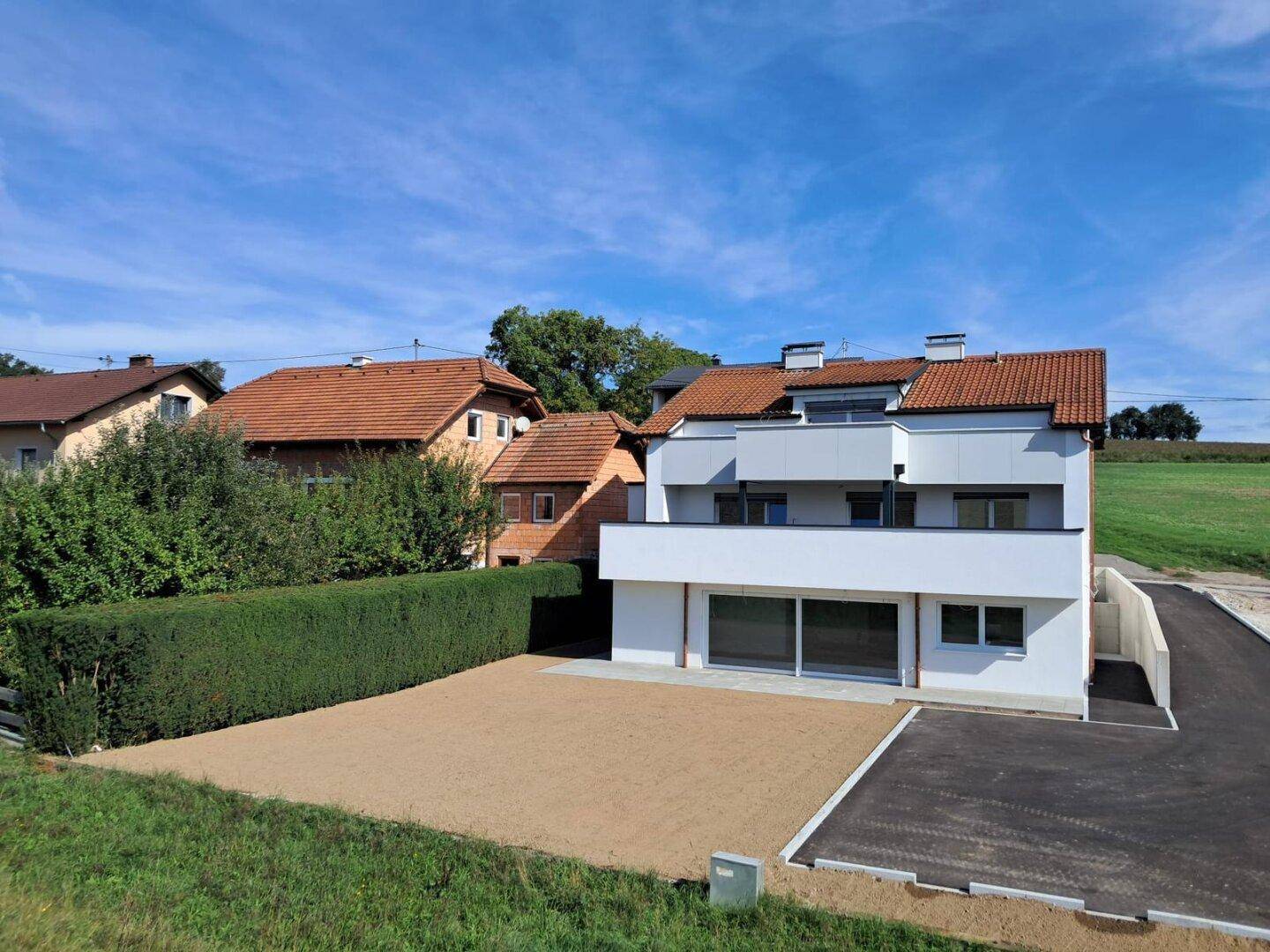 Neubau-Wohnhaus mit moderner Architektur, weißer Fassade und Hecke, eingebettet in ländliche Umgebung.
