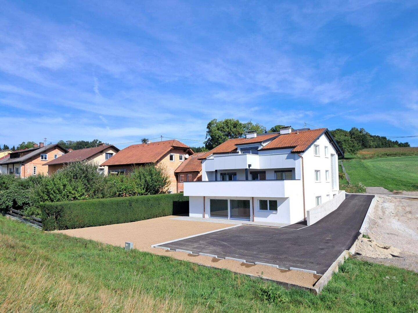 Neues Wohnhaus mit weißer Fassade, rotem Ziegeldach und großzügiger Zufahrt in ländlicher Lage.