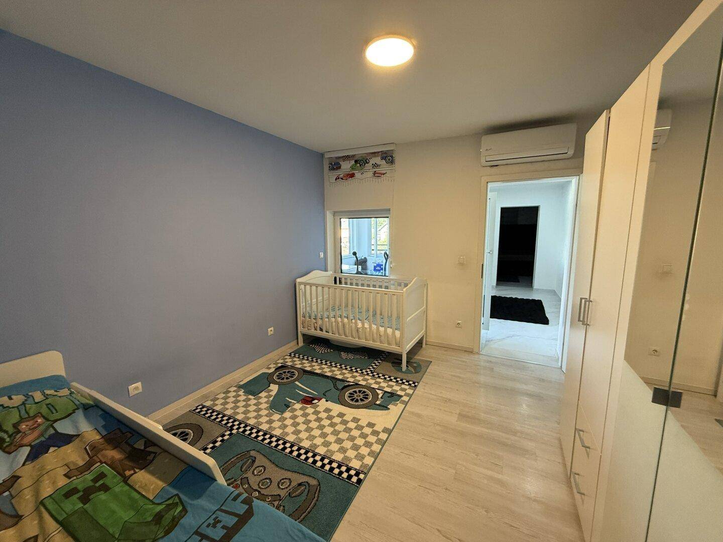 Helles Kinderzimmer mit Babybett, Einzelbett und Klimaanlage.
