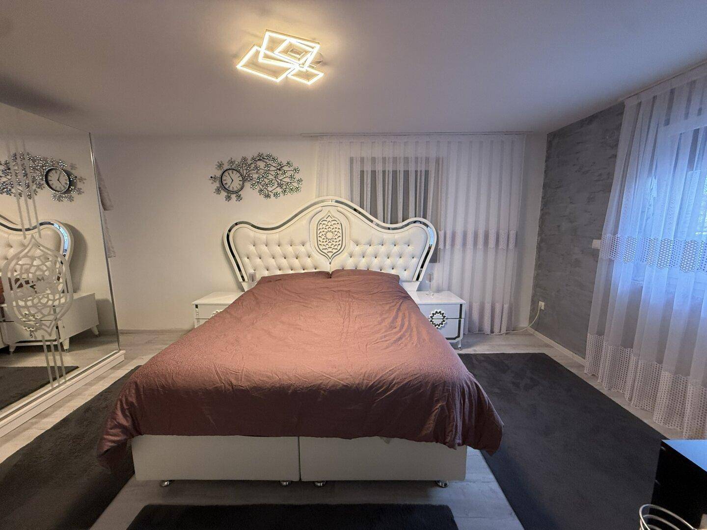 Elegantes Schlafzimmer mit großem, gepolstertem Bett, Spiegel und Vorhängen.